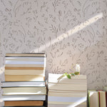 A wall adorned with a delicate floral sketch wallpaper in soft neutral tones, lit by natural sunlight casting a warm glow over a stack of books, evoking a peaceful and scholarly atmosphere.