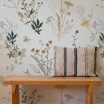 This image features an interior decor scene with the Aerie Floral Wallpaper adorning the wall. The wallpaper displays a serene botanical pattern with soft pastel flowers and foliage. A simple wooden bench with a striped cushion is placed in front of the wallpaper, enhancing the warm and inviting ambiance of the room.