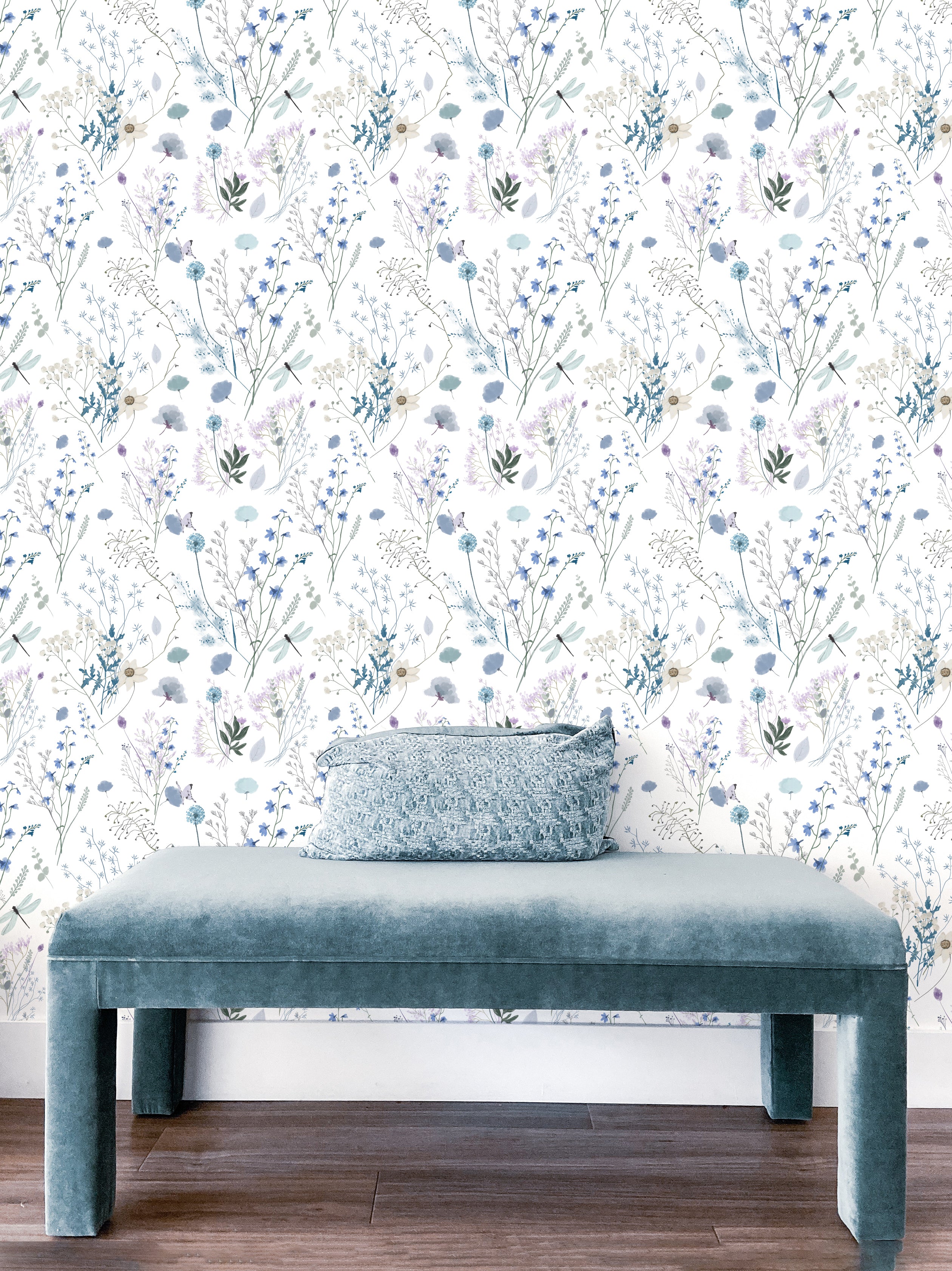 A modern interior space highlighted by the Aerie Floral II Wallpaper with its airy botanical pattern creating a tranquil ambiance. A plush velvet bench in a coordinating blue shade sits against the wallpaper, accented by a textured throw pillow, showcasing a harmonious and stylish setting.