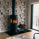 A cozy living room corner enhanced by the Modern Floral Sketch Wallpaper - 25", with black abstract floral patterns on a white background. The wallpaper surrounds a modern, freestanding fireplace, adding a dramatic and stylish element to the warm and inviting room