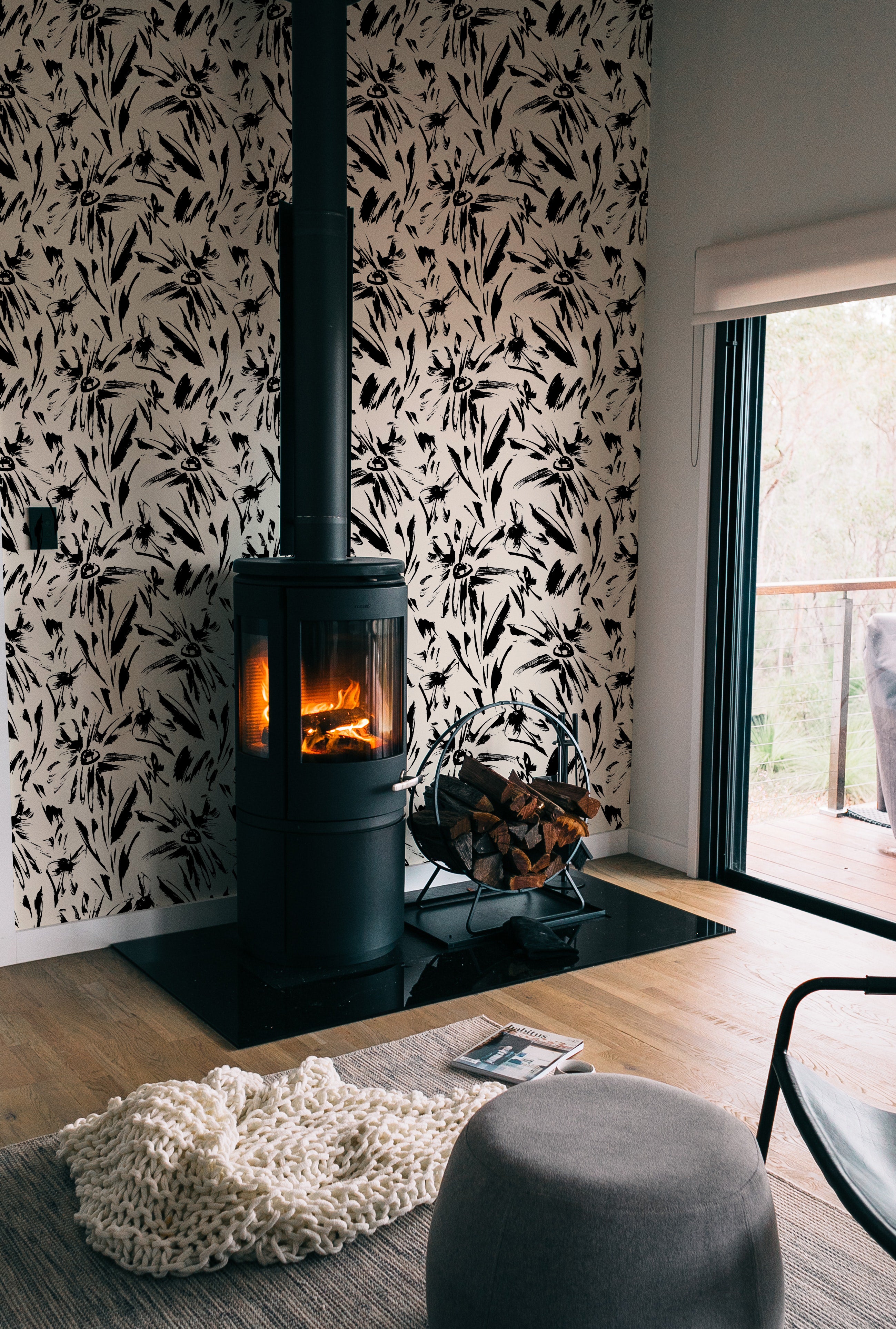 A cozy living room corner enhanced by the Modern Floral Sketch Wallpaper - 25", with black abstract floral patterns on a white background. The wallpaper surrounds a modern, freestanding fireplace, adding a dramatic and stylish element to the warm and inviting room