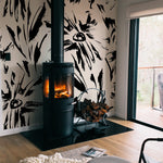 A cozy room setup showcasing the Modern Floral Sketch Wallpaper - 75" around a freestanding modern fireplace. The bold, black abstract floral patterns contrast strikingly with the white background, adding a dramatic flair to the warm and inviting space