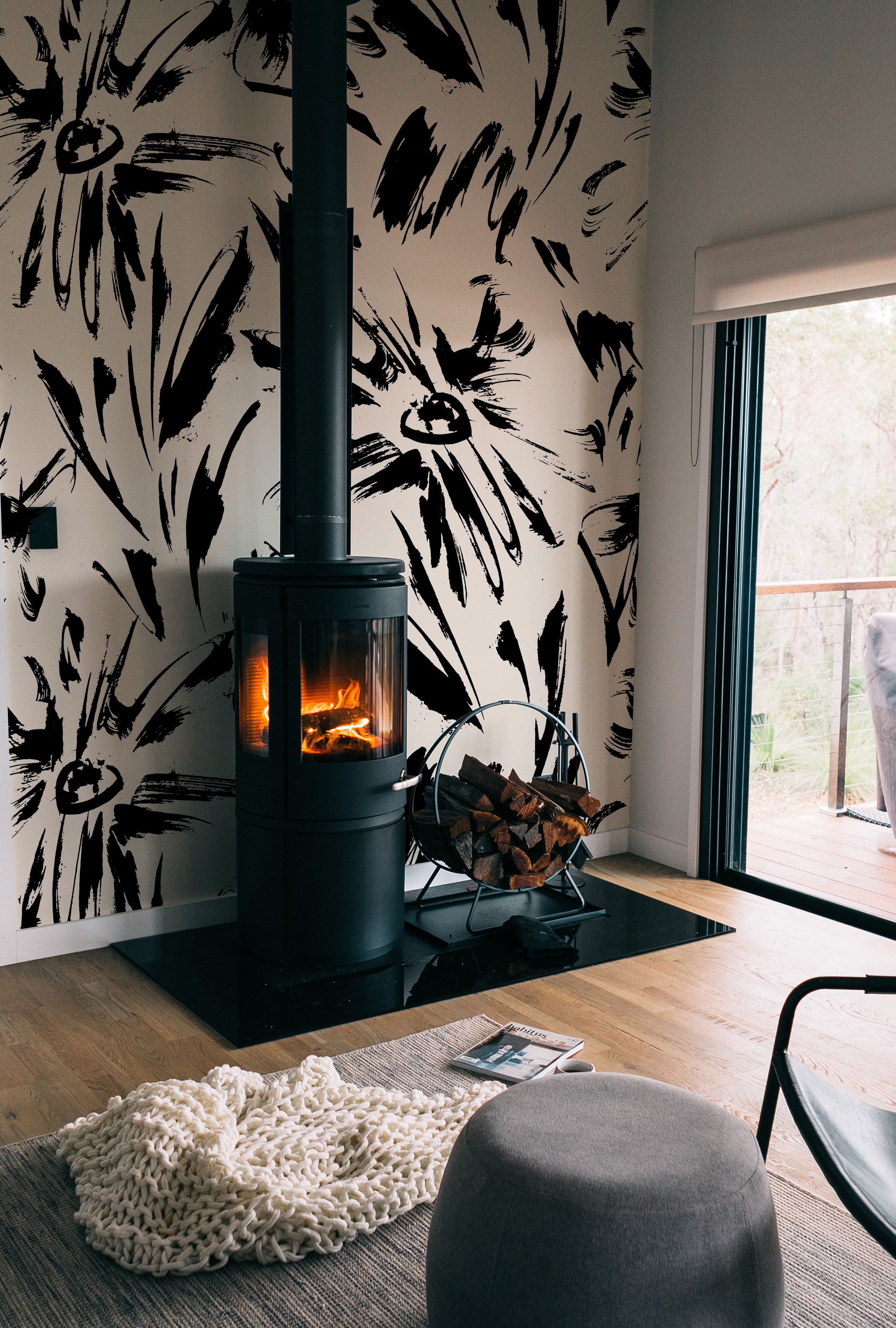 A cozy room setup showcasing the Modern Floral Sketch Wallpaper - 75" around a freestanding modern fireplace. The bold, black abstract floral patterns contrast strikingly with the white background, adding a dramatic flair to the warm and inviting space