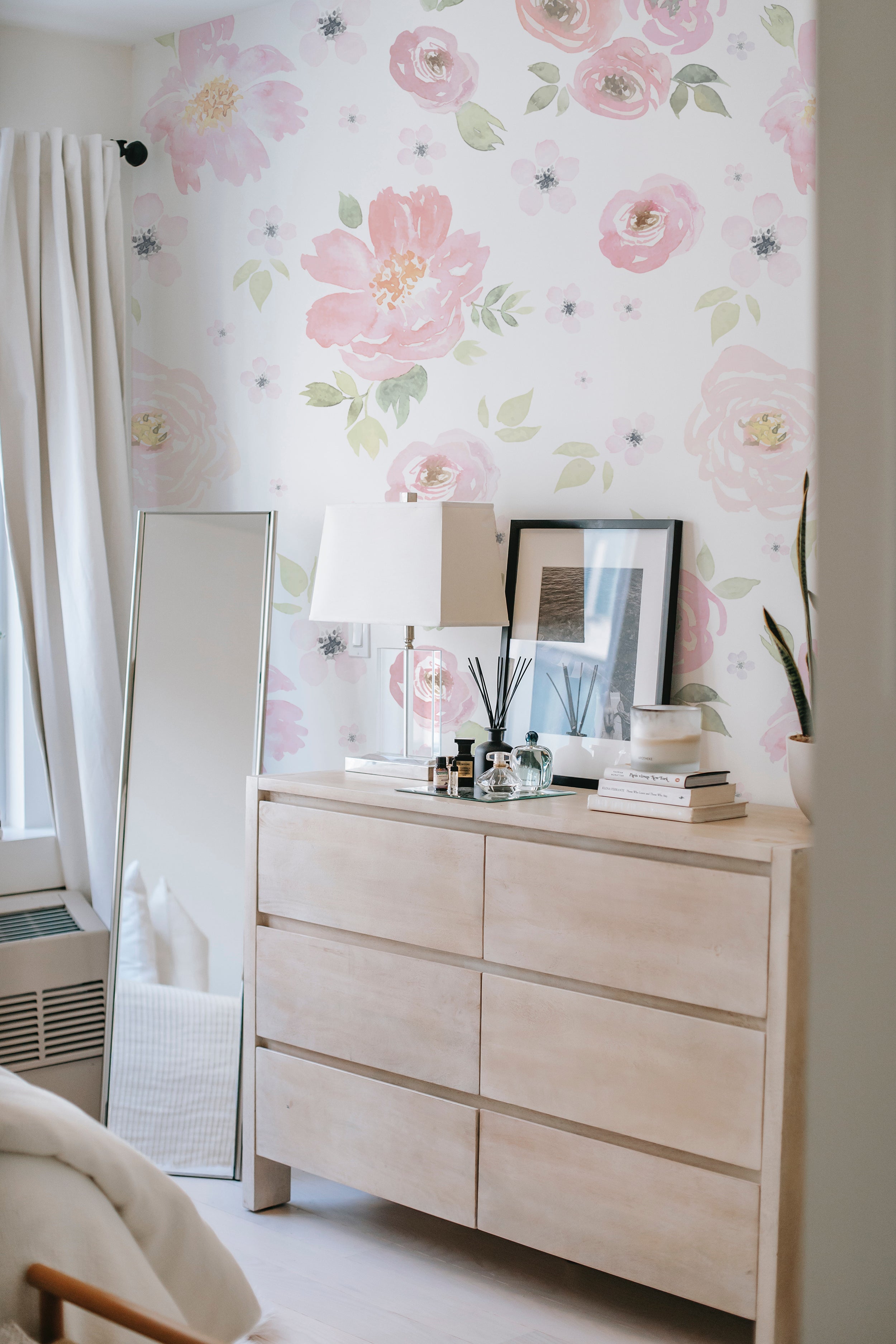 A stylish bedroom setup showcasing the Gentle Blossom Wallpaper - 75" on the main wall behind a contemporary wooden dresser and a large mirror. The wallpaper's lush pink floral pattern adds a touch of femininity and warmth, complementing the minimalistic decor and neutral furniture