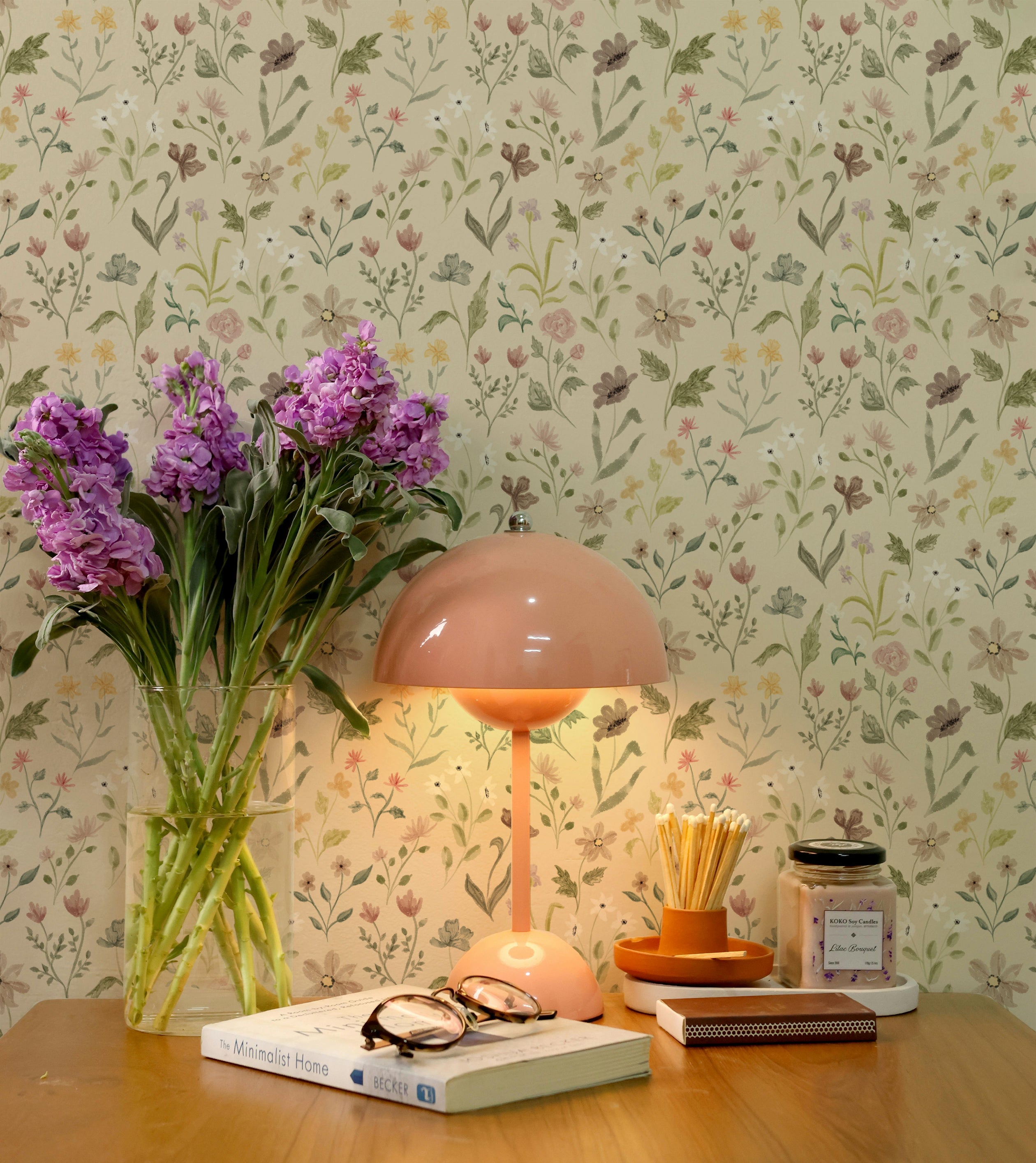 A wooden desk with a pink lamp, vase of purple flowers, books, and a candle, complemented by the Whimsy Wildflowers Wallpaper, featuring a cheerful and colorful floral design in pastels.
