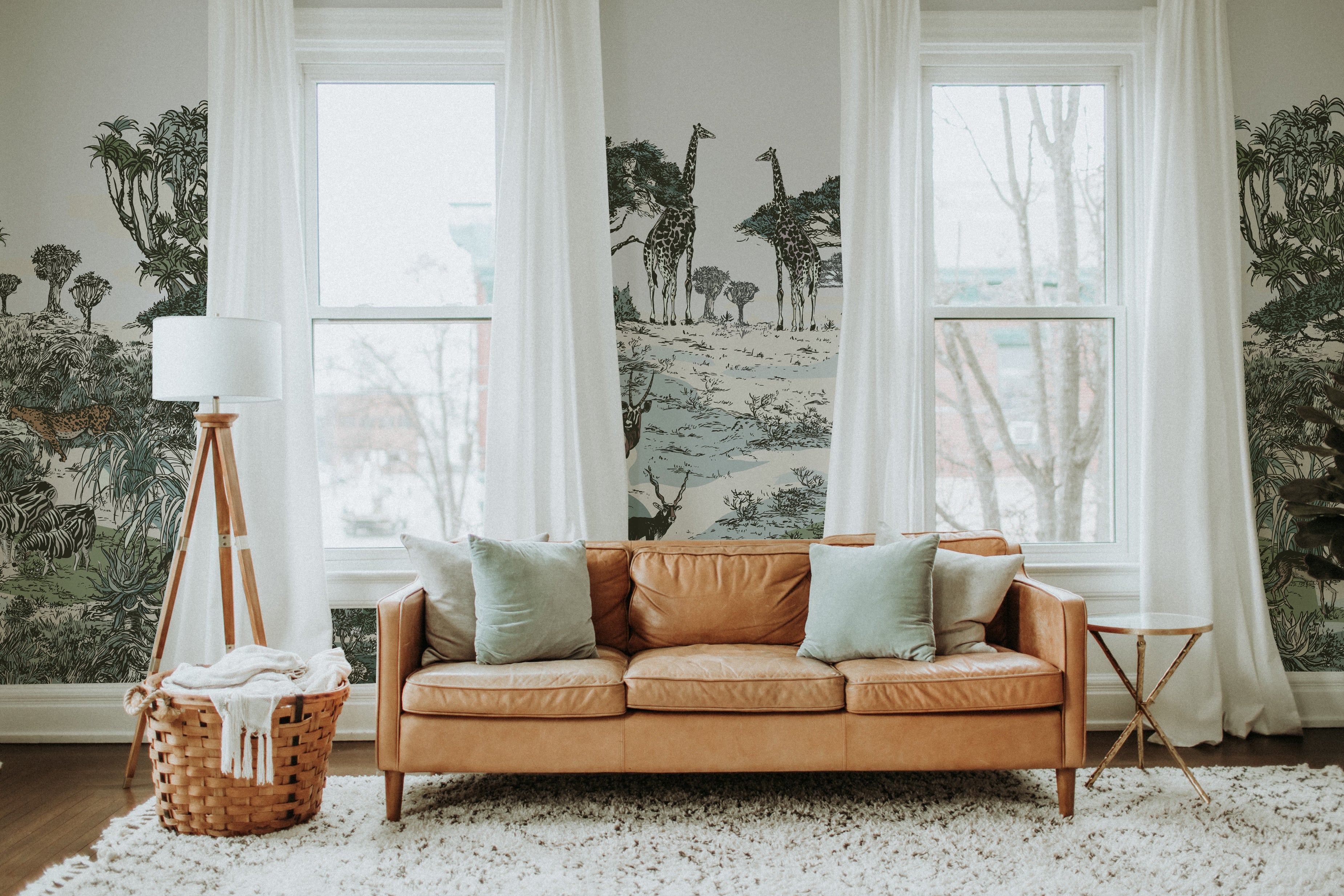 A bright living room space with large windows draped in sheer white curtains. A caramel leather couch with mint green pillows sits against a wall adorned with safari-themed wallpaper featuring giraffes and lush vegetation