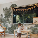 A child's play area with a safari wallpaper backdrop. A small wooden table and stools are set for play, and a rattan pouffe is nearby. Black and white bunting hangs above the window, and wooden play bowls are arranged on the floor