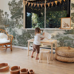 A child's play area with a safari wallpaper backdrop. A small wooden table and stools are set for play, and a rattan pouffe is nearby. Black and white bunting hangs above the window, and wooden play bowls are arranged on the floor