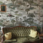 A vintage green velvet tufted sofa with two mustard yellow throw pillows. A rustic wooden side table with books and a potted plant is placed to the right of the sofa. The interior is complemented by safari-inspired wallpaper featuring a savanna landscape with trees, impalas, and a leopard. An ornate framed mirror hangs on the left wall