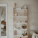 A cozy corner of a room showing Boardwalk Wallpaper enhancing the wall behind a wooden shelving unit. The decor includes various decorative items like plants, books, and ceramics that complement the neutral tones of the wallpaper.