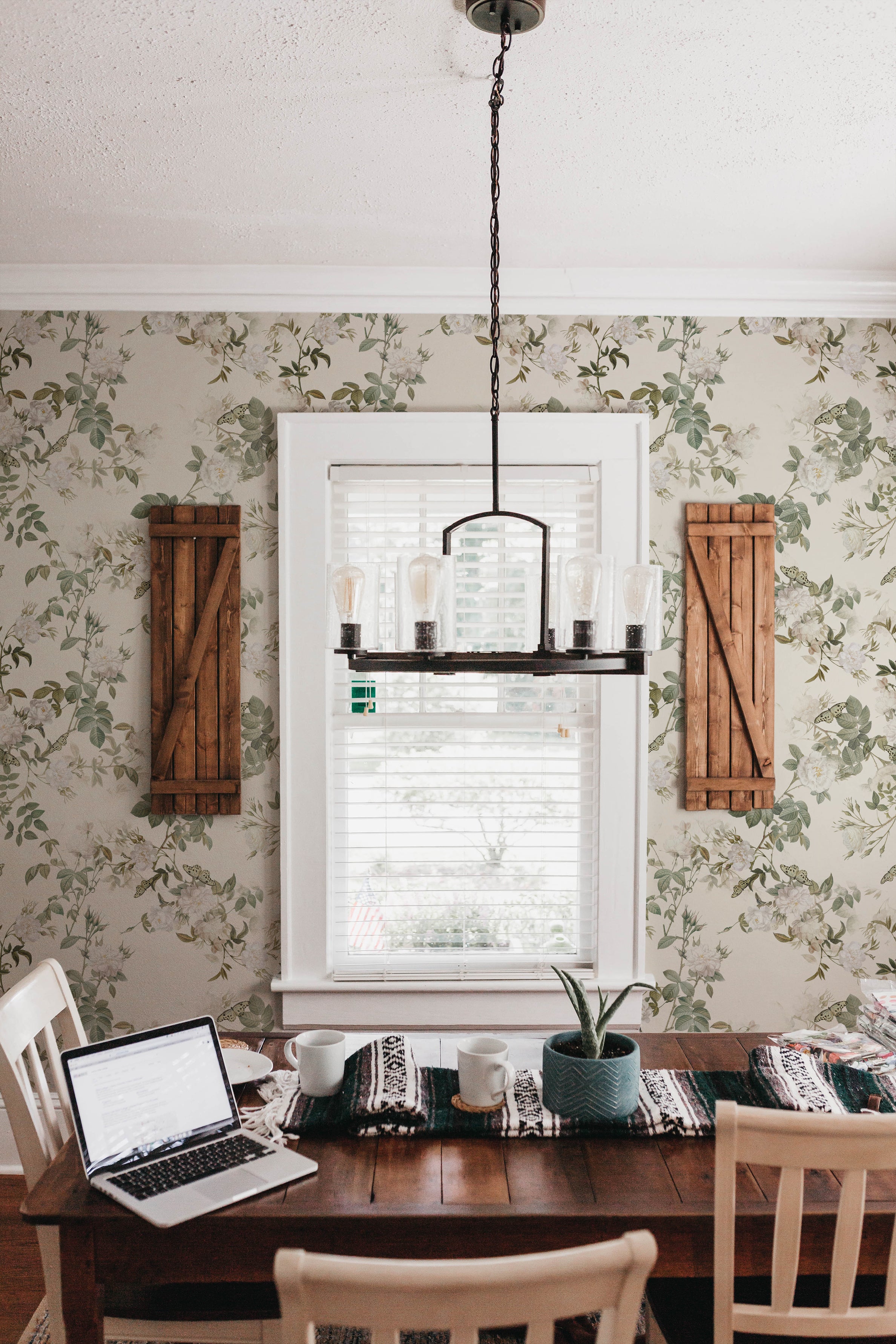An elegant dining room with botanical wallpaper featuring large mint-green leaves and white flowers, a wooden table with a laptop and two mugs, wooden chairs, and a chandelier.
