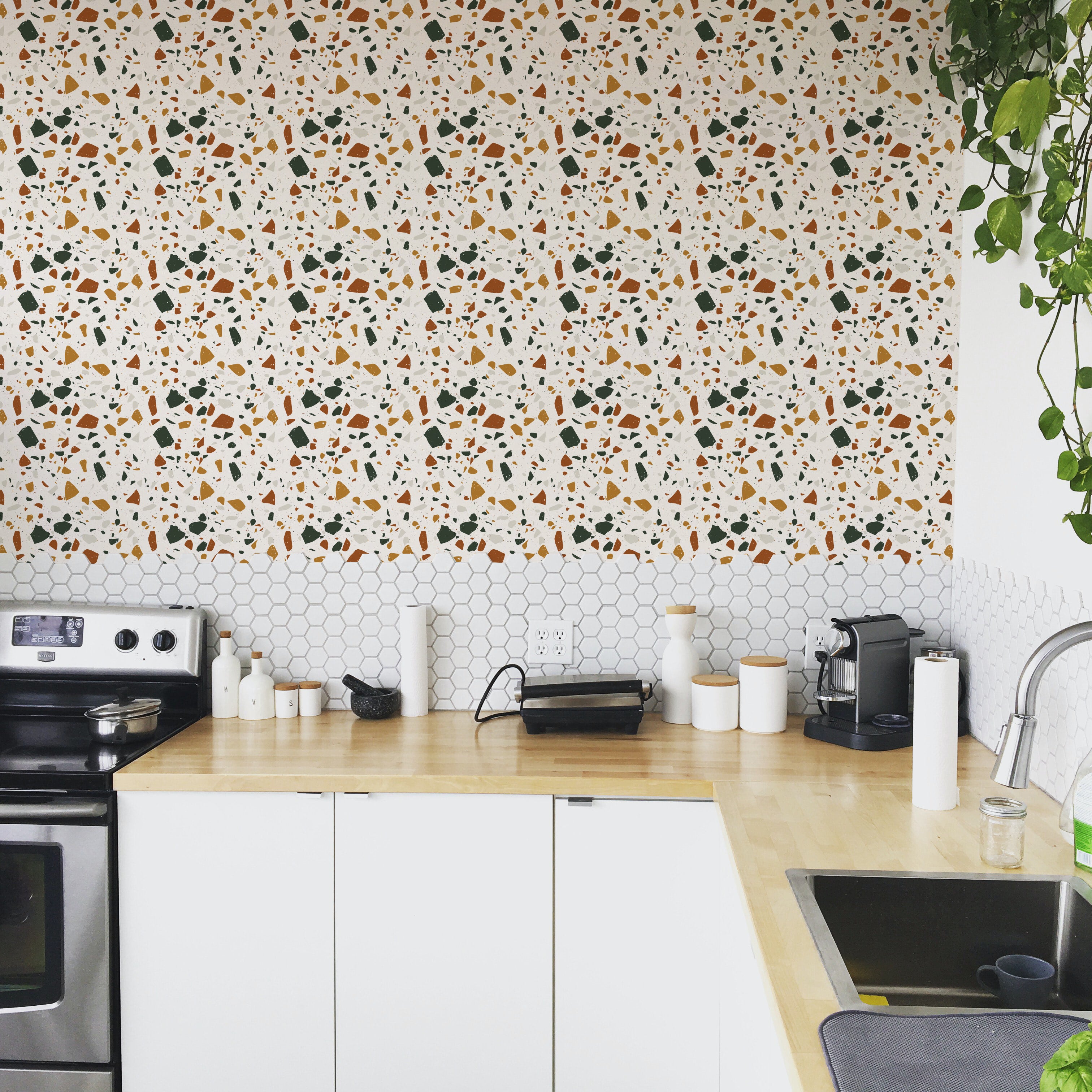 A kitchen backsplash covered with Earthy Terrazzo wallpaper, showing a vibrant and colorful pattern of orange, green, and gray specks on a white background, complemented by white cabinetry and modern kitchen appliances.