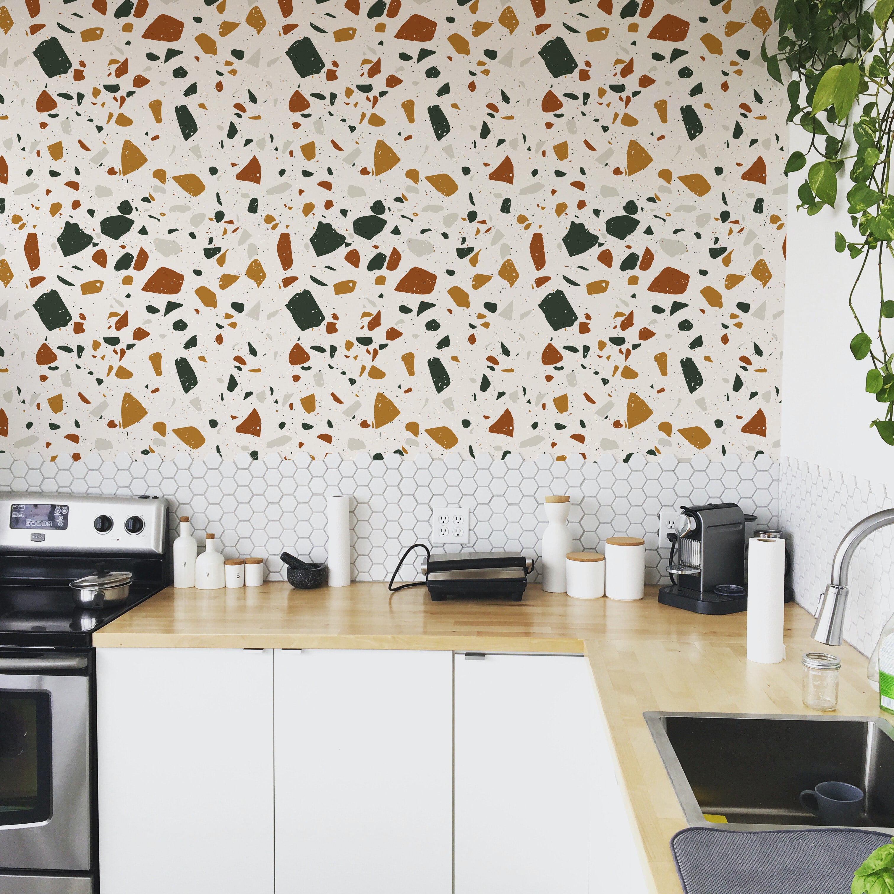 A stylish kitchen featuring a backsplash adorned with Earthy Terrazzo wallpaper, showcasing a colorful terrazzo pattern with orange, green, and gray chips on a white background, complementing white kitchen cabinets and hexagonal tiles.