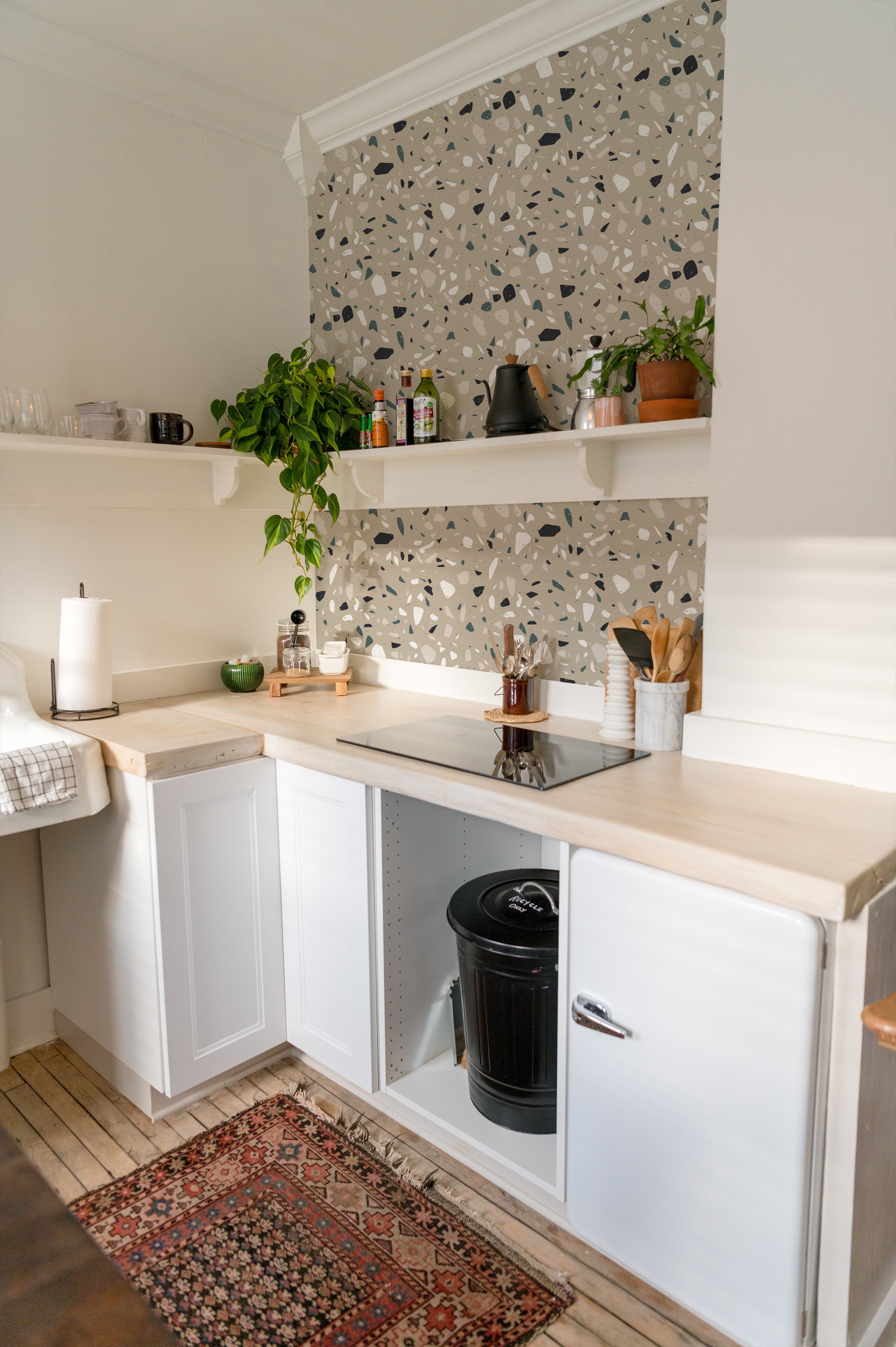 A kitchen setting featuring the Earthen Terrazzo Wallpaper as a backsplash, enhancing the space with its stylish terrazzo pattern of irregular shapes in muted tones. The design complements the white cabinetry and natural wood countertops, creating a modern yet earthy ambiance.