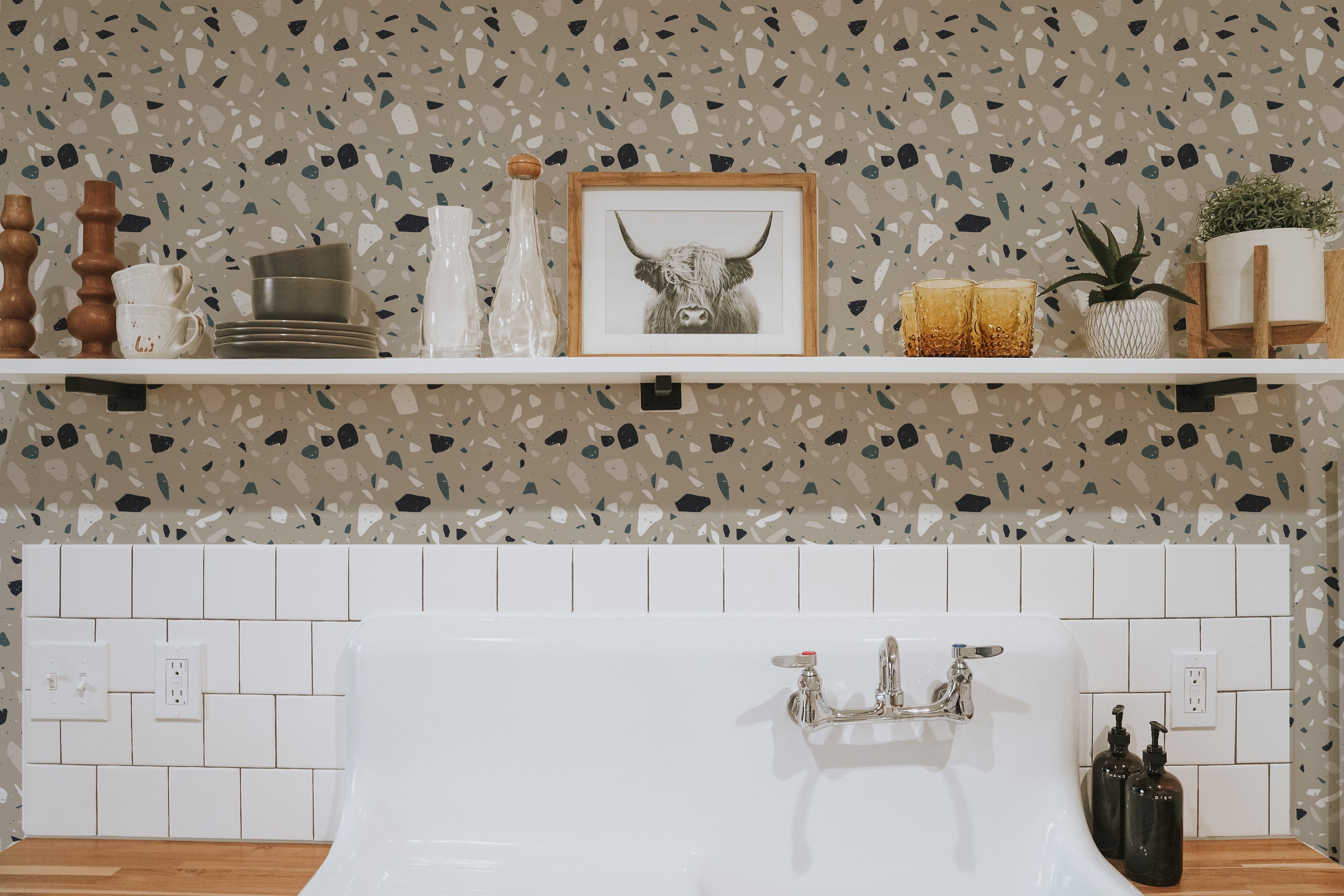 A bathroom setting featuring the Earthen Terrazzo Wallpaper as a backsplash, enhancing the space with its stylish terrazzo pattern of irregular shapes in muted tones. The design complements the white cabinetry and natural wood countertops, creating a modern yet earthy ambiance.