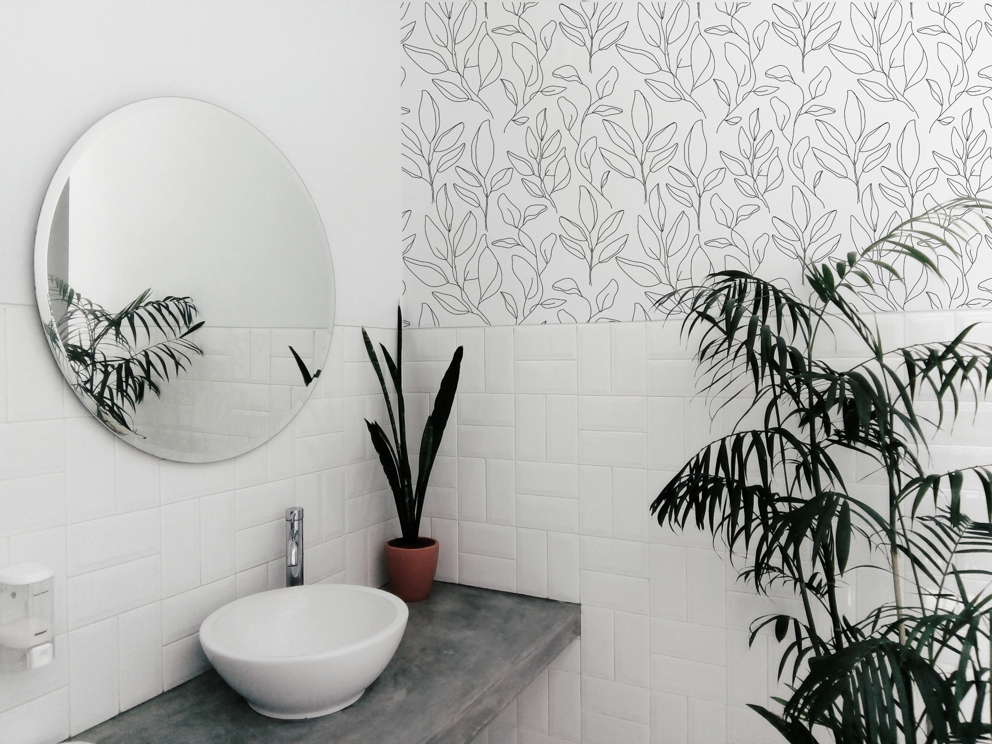 A contemporary bathroom featuring a round mirror, white sink, and green plants. The upper part of the wall is decorated with Black Floral Wallpaper, featuring a delicate black line art design of leaves on a white background.