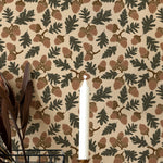 Interior wall decorated with a seamless botanical wallpaper pattern featuring acorns and oak leaves in earthy tones. A wooden sideboard with vertical lines stands against the wall, adorned with a wire basket holding feathers and a brass candlestick with a single white candle