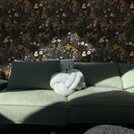 A chic living room with sunlight streaming across a plush L-shaped sofa adorned with a unique braided cushion. The wall behind features a dark botanical wallpaper with a lush pattern of white and mustard flowers set against a backdrop of dark leaves