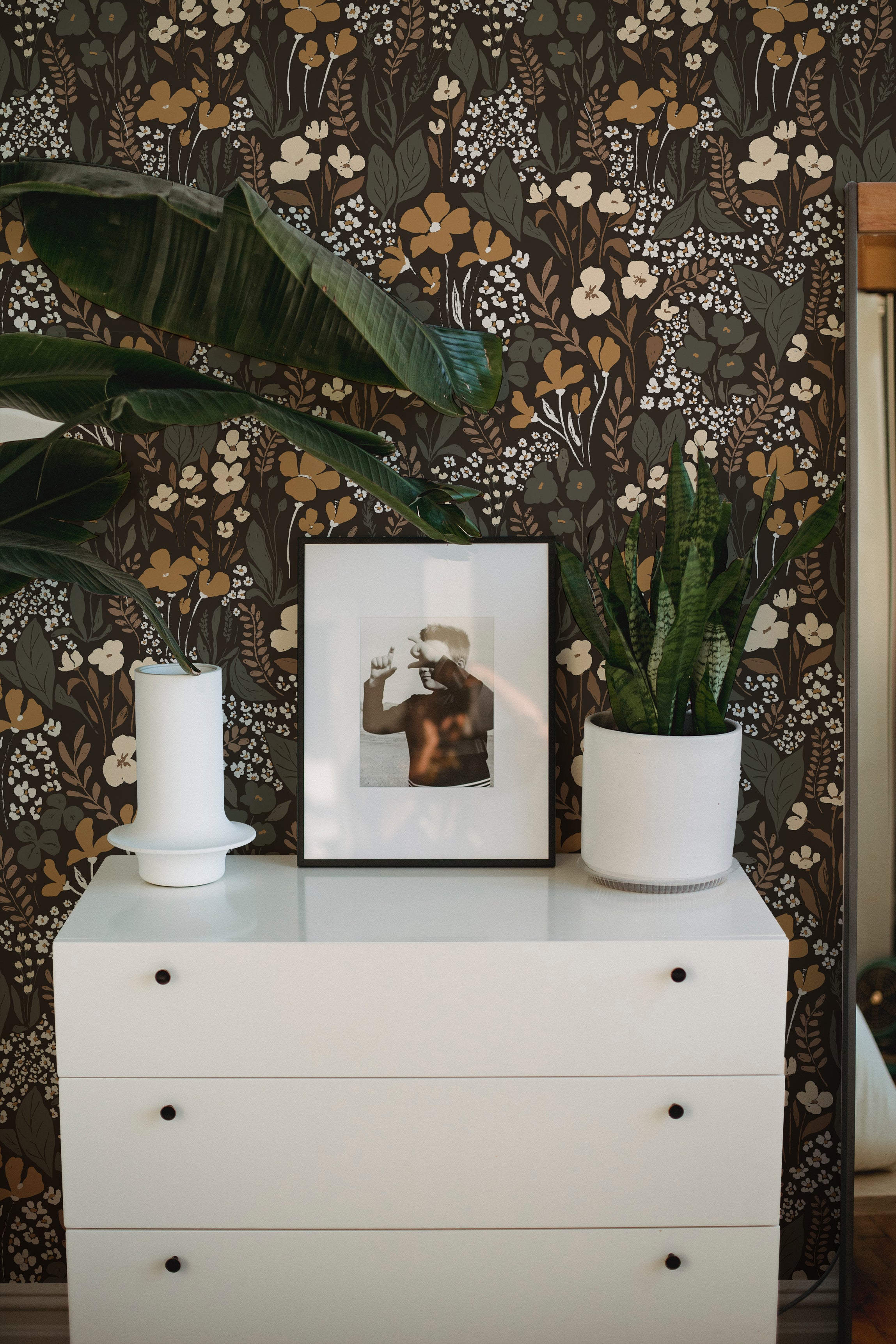 A modern dresser in white with a minimalistic design set against a botanical wallpaper backdrop with a dense floral pattern in dark brown, white, mustard, and green hues. The dresser is styled with a white candlestick, a framed photograph, and a potted snake plant, enhancing the room's organic elegance