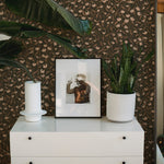 Elegant and modern interior with a white dresser set against a backdrop of sophisticated dark floral wallpaper, featuring small, delicate light pink flowers and brown leaves, complemented by green houseplants, a framed photograph, and a white candle.