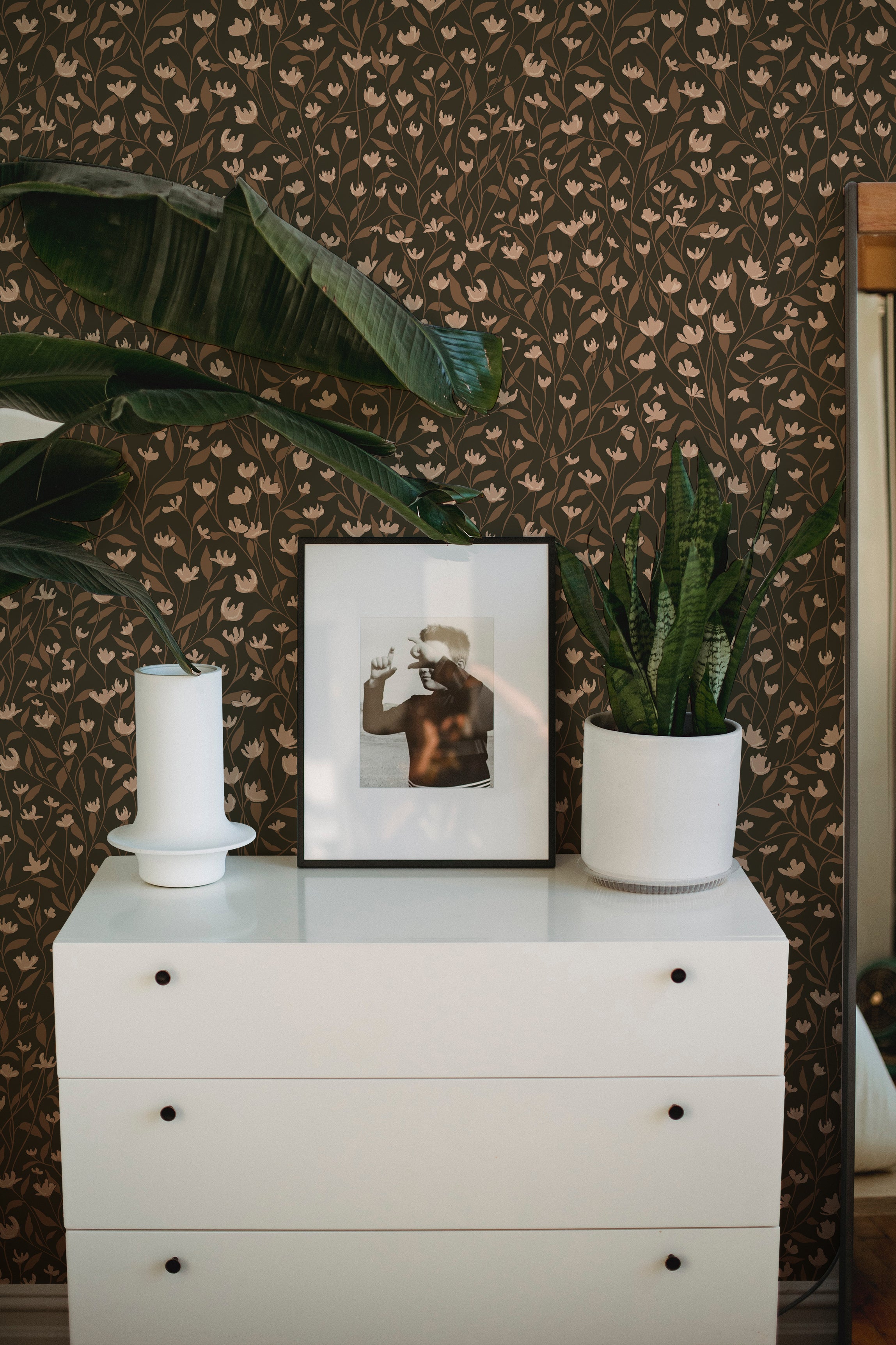 Elegant and modern interior with a white dresser set against a backdrop of sophisticated dark floral wallpaper, featuring small, delicate light pink flowers and brown leaves, complemented by green houseplants, a framed photograph, and a white candle.