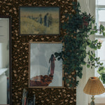 Bohemian-inspired workspace with an ornate dark floral wallpaper providing a striking background to vintage paintings, a potted hanging plant, and a classic table lamp, creating a layered, textured look.