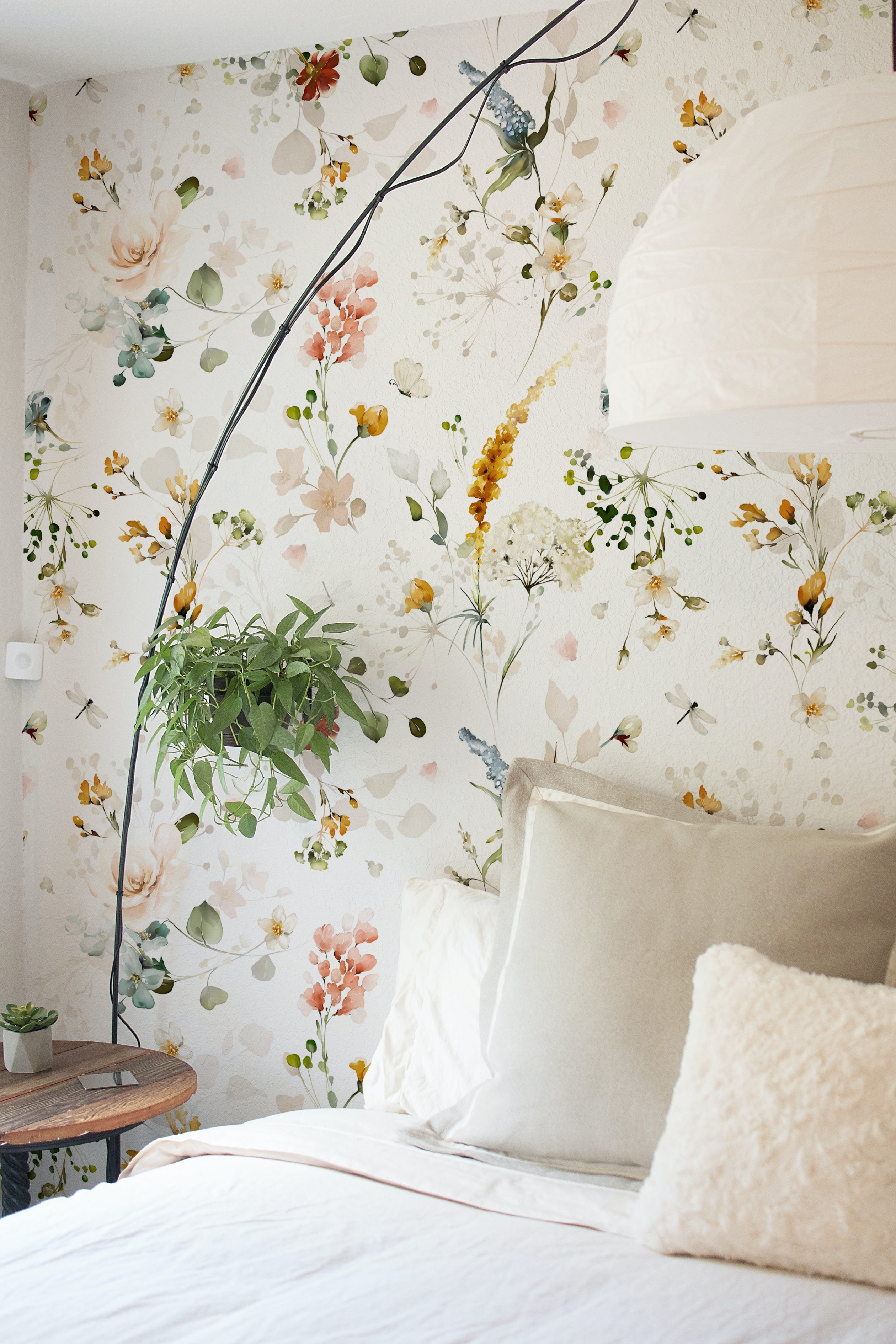 A beautifully styled bedroom featuring Fiori Wallpaper, with a floral pattern that includes lush, watercolor flowers in soft pastel shades adorning the wall. The room includes beige pillows and a white bedspread, enhancing the room's serene and inviting atmosphere.