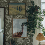 A sophisticated and artistic home office corner featuring an intricate owl-themed wallpaper in black and white, providing a striking backdrop to framed pictures and a lush potted plant