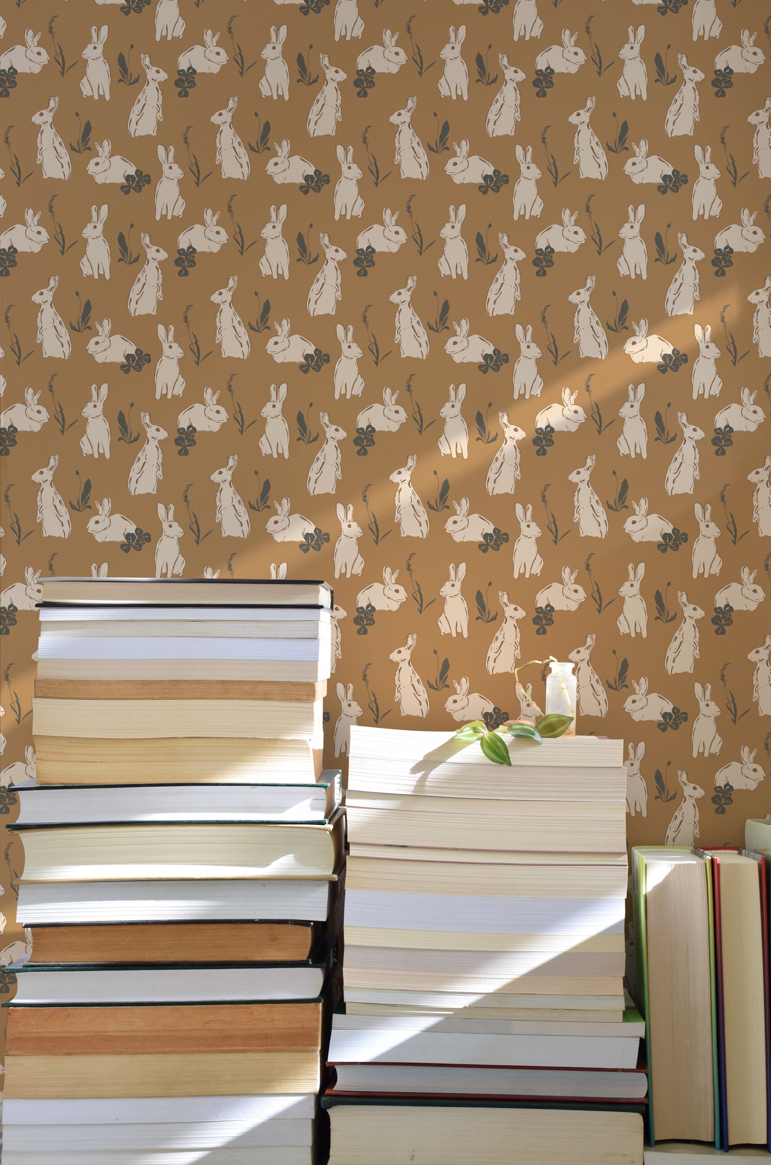 A close-up of charming rabbit-patterned wallpaper, with sketched rabbits and flora in beige and cream, complemented by the warm glow of sunlight across a stack of books