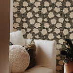 A cozy corner with a white armchair and a crochet footstool against a wall with a large-scale floral wallpaper pattern, featuring stylized roses in cream on a dark background with olive green foliage.