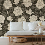 Chic, minimalist lounge area adorned with a floral wallpaper showcasing large cream roses on a dark background. The setting includes a light gray sofa, a natural wood coffee table, and a woven floor pouf, emphasizing a clean and tranquil space