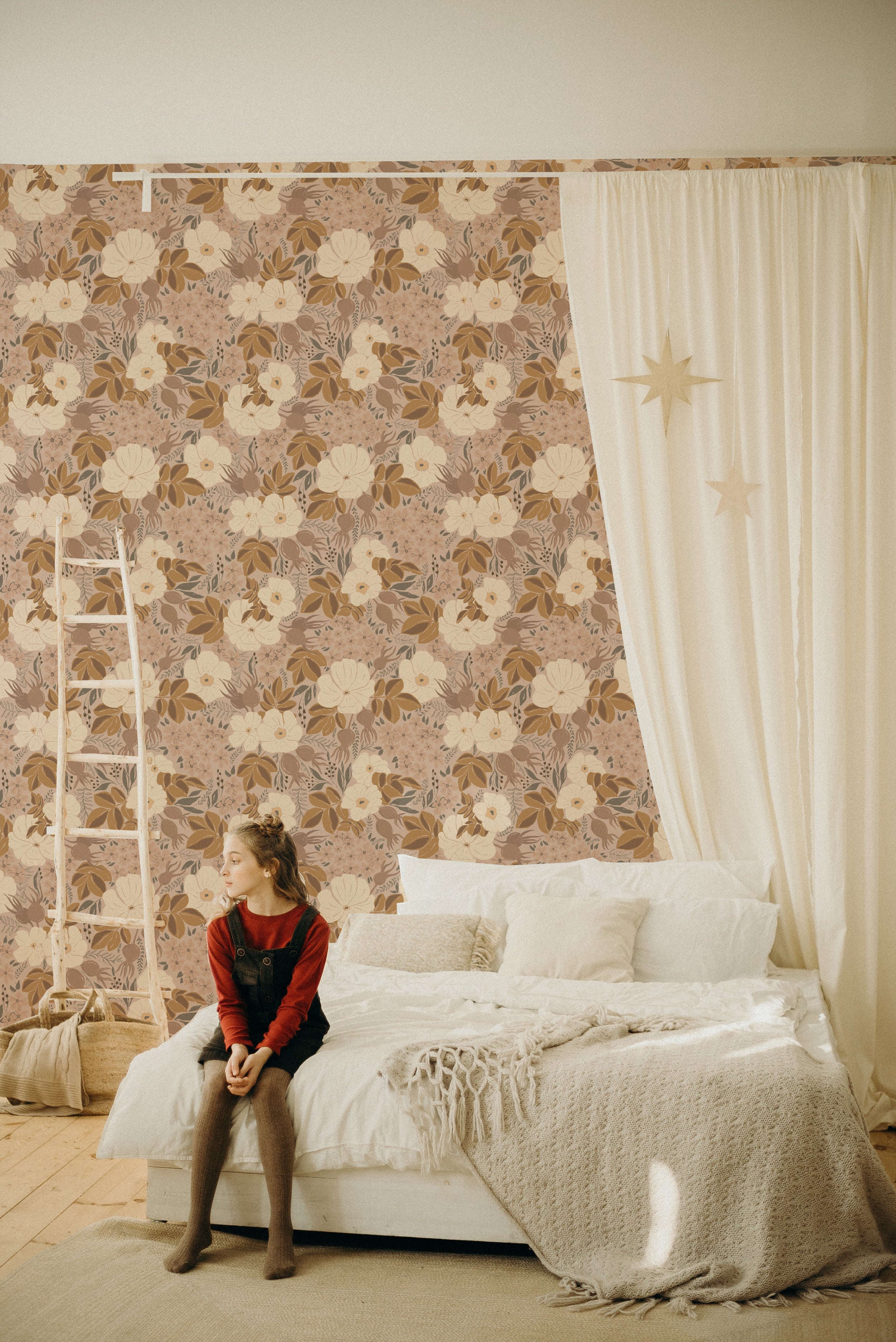 A cozy bedroom featuring a wooden ladder, a large bed with white bedding, and a light curtain adorned with star patterns. The room is decorated with the Rosehip Wallpaper, which displays a rich floral pattern with large white blooms and smaller pink flowers against a dark backdrop, adding a warm and inviting atmosphere.