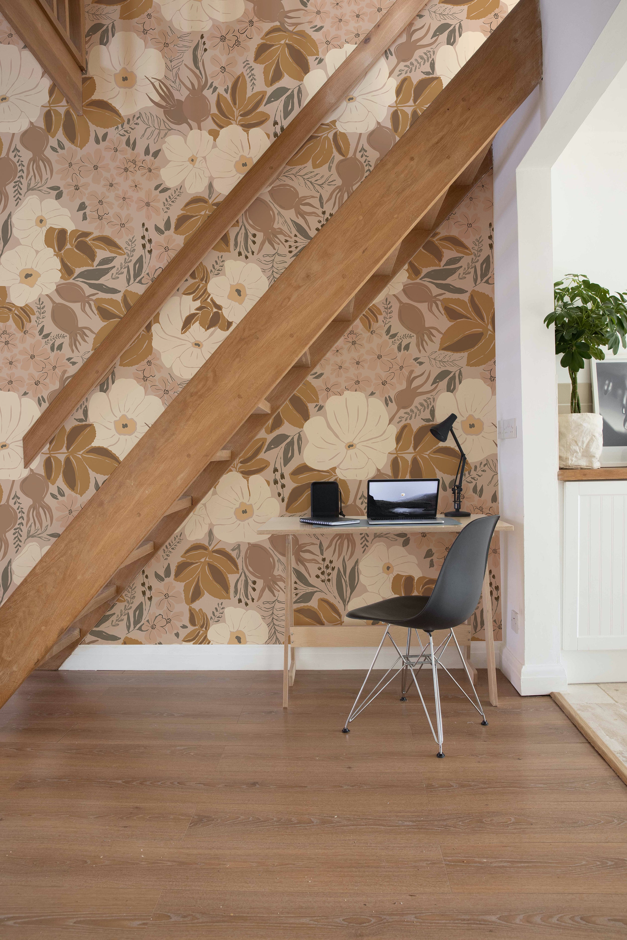 A compact home office situated under a staircase, with a large floral wallpaper providing a vibrant backdrop. The workspace includes a simple desk, a modern chair, and a small laptop