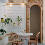 A quaint café corner decorated with a large floral wallpaper in earthy hues. The space includes a small round table, curved wooden chairs, and a series of potted plants on white floating shelves.