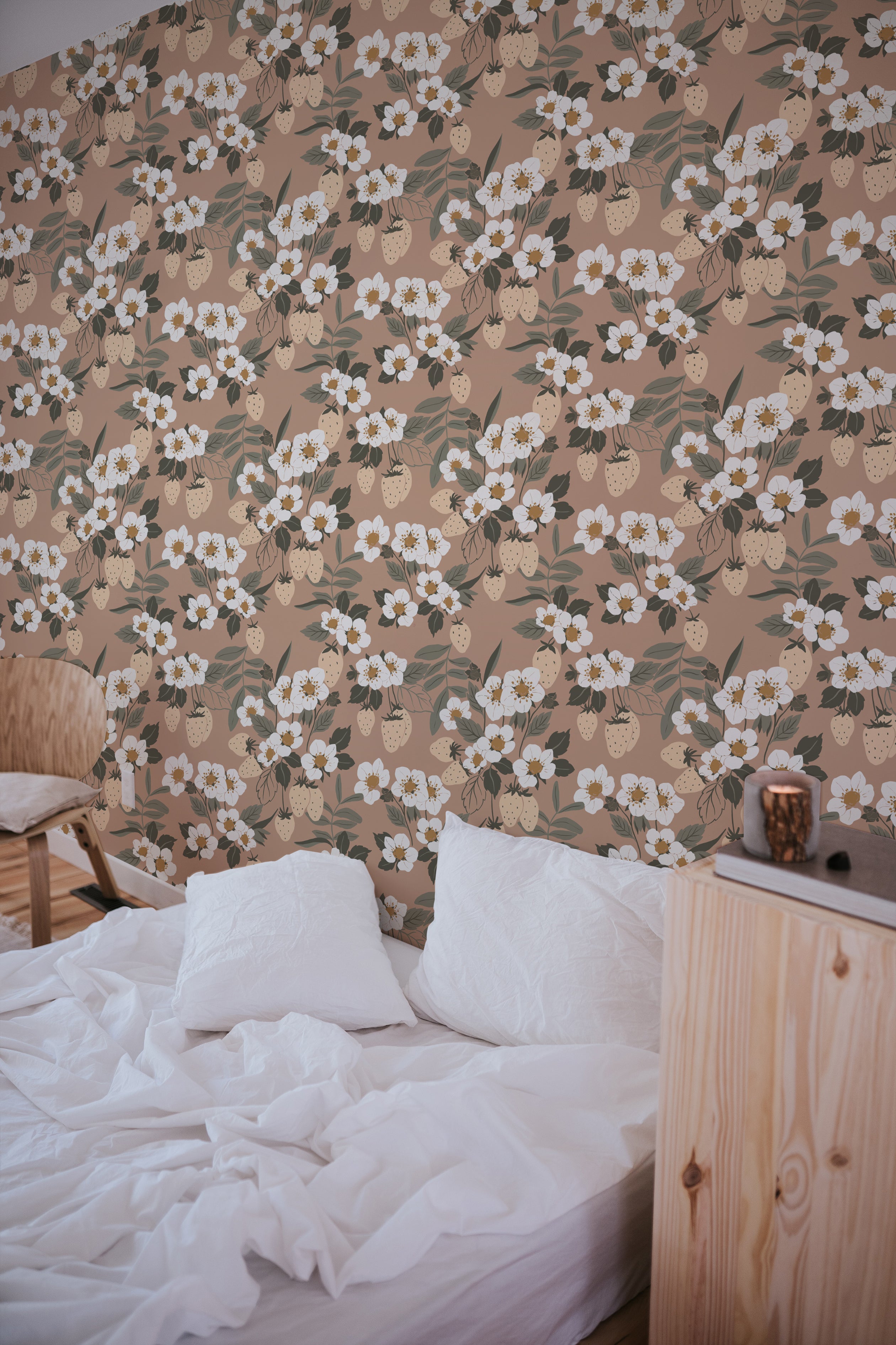 A cozy bedroom scene featuring a simple wooden bed with white bedding, set against a background of Forest Strawberries Wallpaper. The wallpaper has a charming pattern of white flowers and light green leaves interspersed with soft pink strawberries on a warm taupe background.
