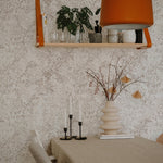 A cozy dining room scene with walls adorned in the Golden Delicate Winter Floral wallpaper, complementing the earth-toned furnishings and decor, including a beige tablecloth, wooden shelf with plants, and a vibrant orange lampshade.