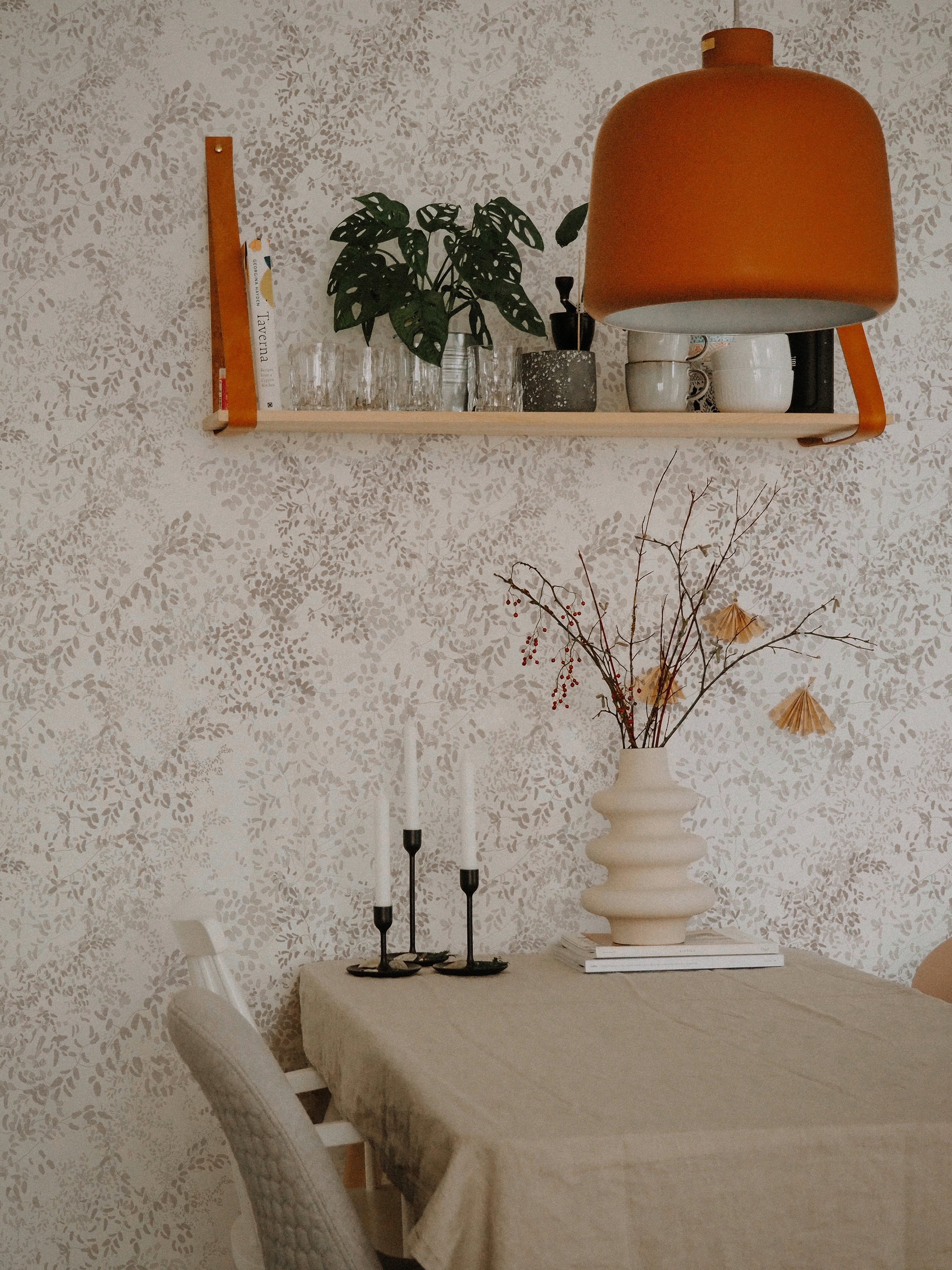 A cozy dining room scene with walls adorned in the Golden Delicate Winter Floral wallpaper, complementing the earth-toned furnishings and decor, including a beige tablecloth, wooden shelf with plants, and a vibrant orange lampshade.