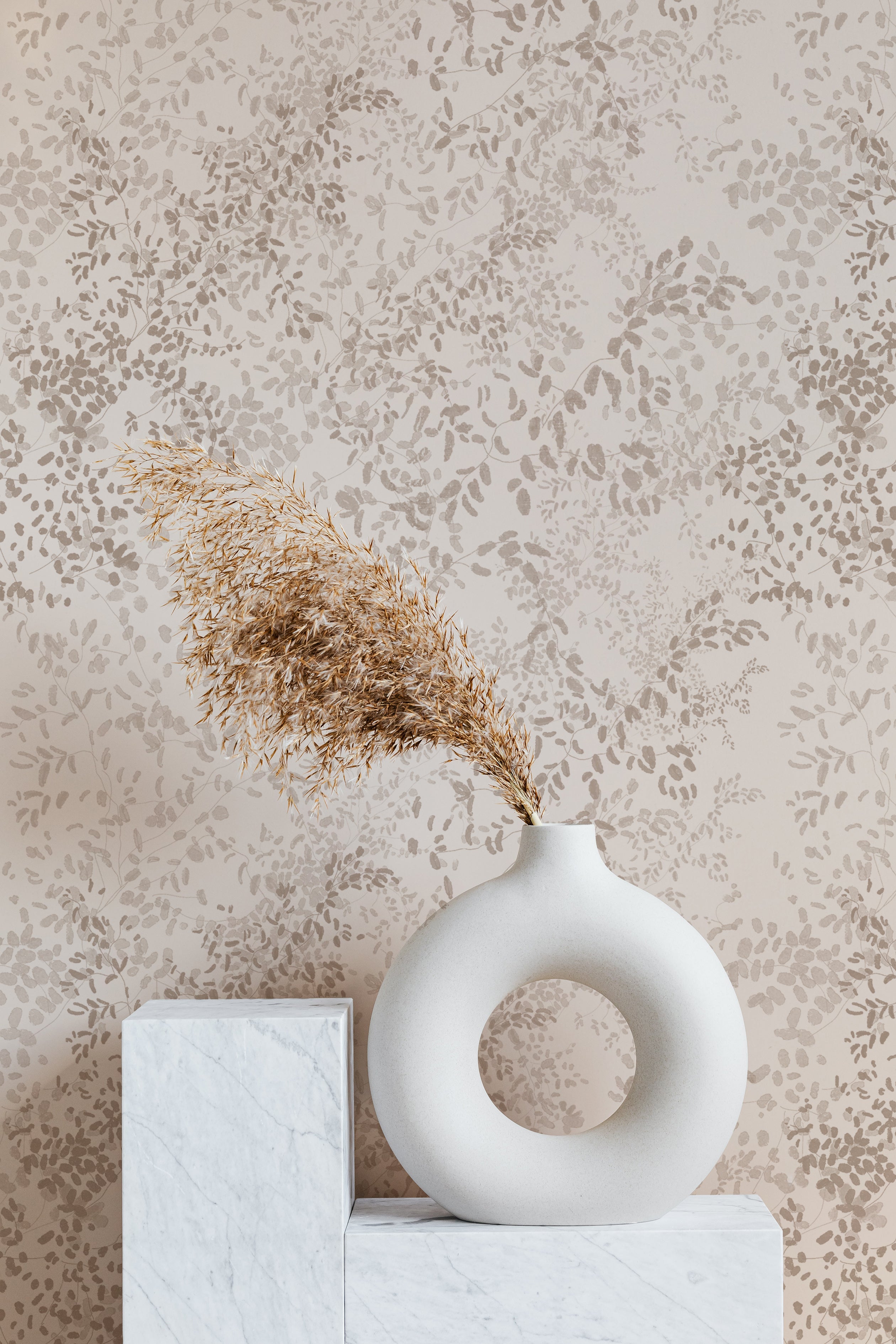 A modern interior setting featuring the Golden Delicate Winter Floral wallpaper as a backdrop with a white vase holding golden dried grass on a marble and white pedestal, blending effortlessly with the subtle floral design.