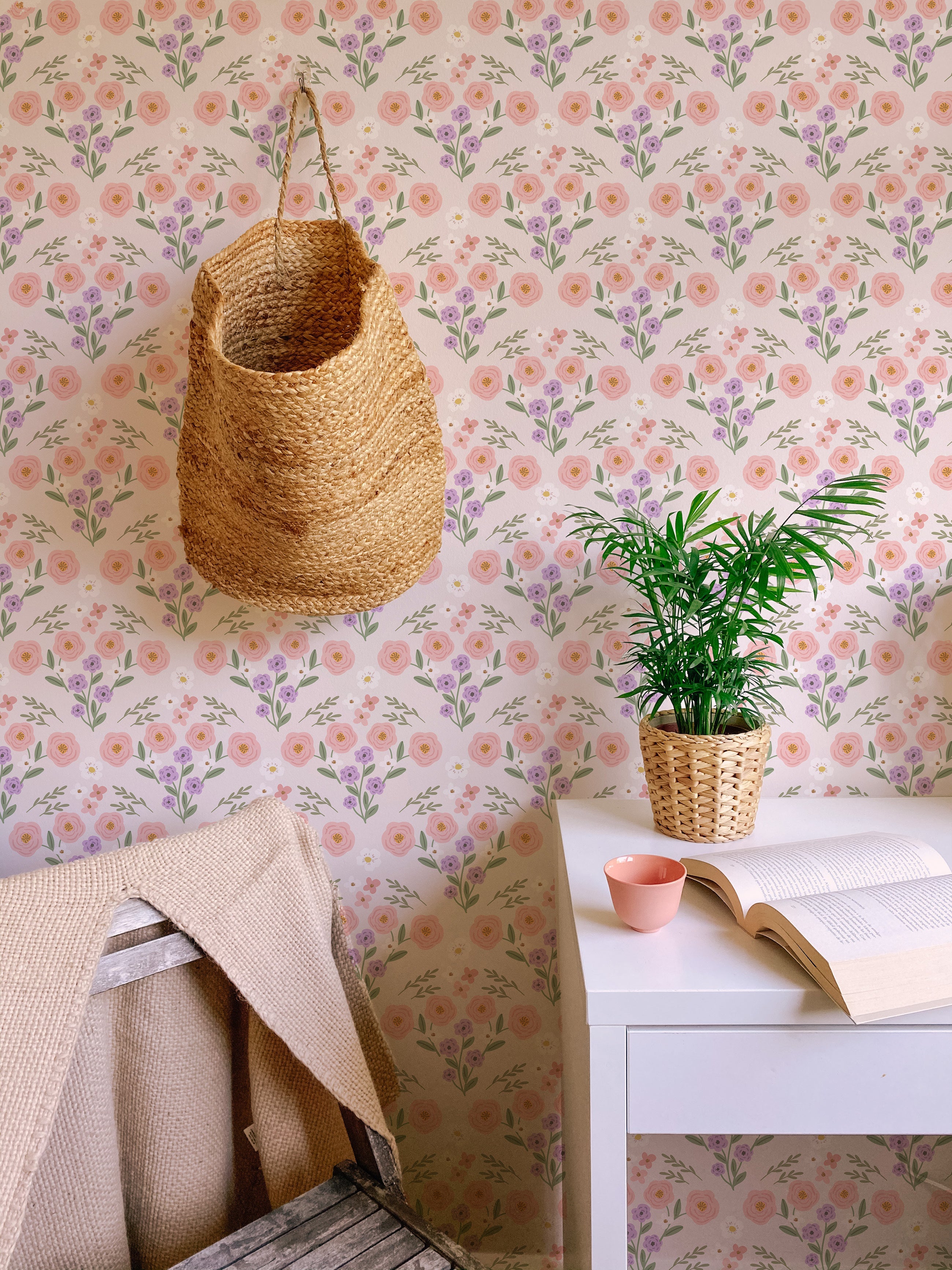 A cozy reading corner enhanced by Fredlig Flowers Wallpaper, which covers the walls with its cheerful pattern of pink roses and purple flowers. A natural wicker basket hangs from the ceiling, complementing the botanical theme alongside a potted green plant and a book on a white desk, providing a peaceful retreat.