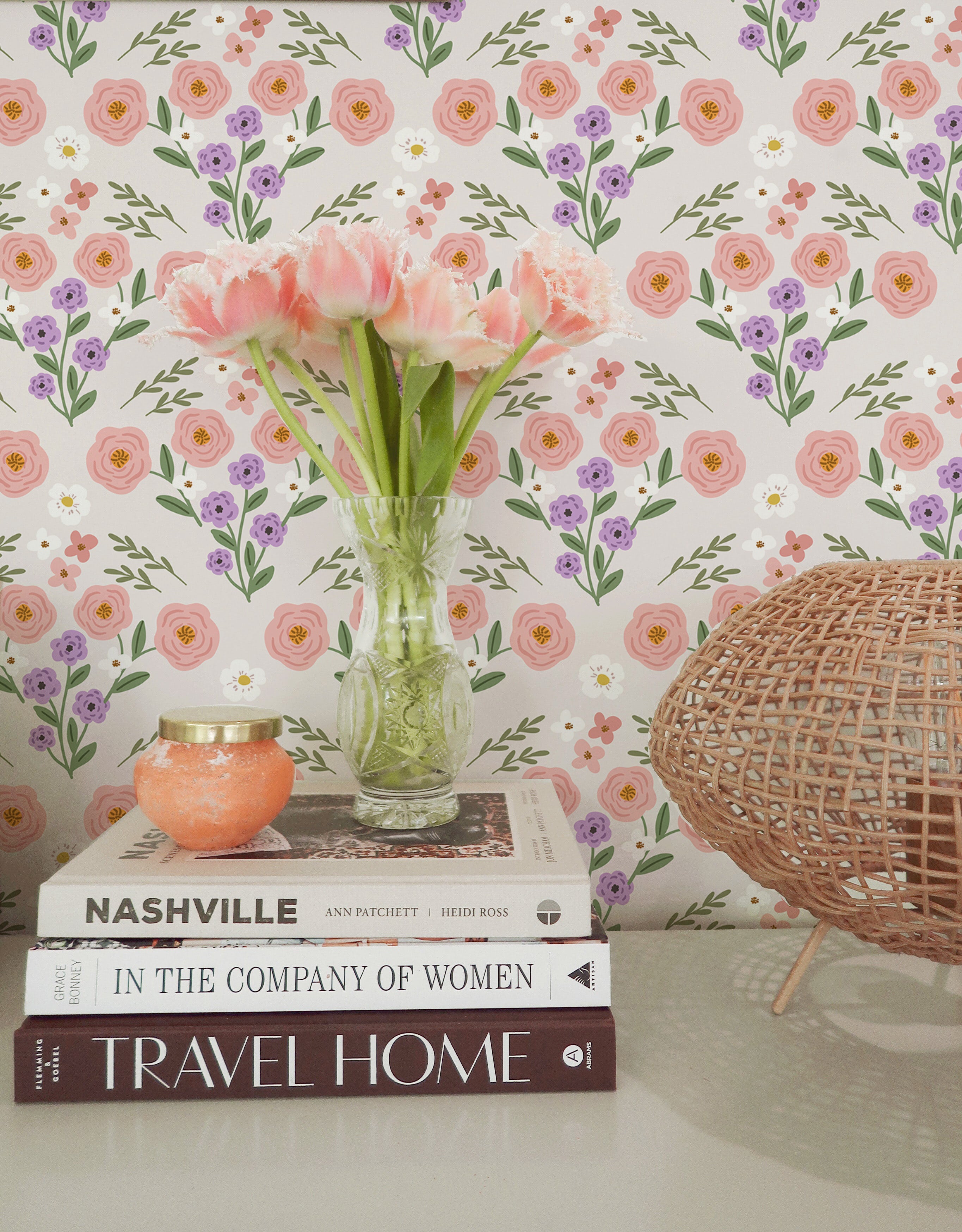 A charming home decor scene featuring Fredlig Flowers Wallpaper, adorned with a floral pattern of pink and purple blooms set against a white background. The arrangement includes a vase with pink tulips, a woven basket, and a stack of books, creating a warm and inviting atmosphere.