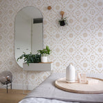 A cozy bedroom setting enhanced by Euclid Wallpaper, which covers the wall behind a bed and a small shelf with a decorative mirror and green houseplants. The wallpaper's soft beige geometric patterns on white provide a calm and inviting backdrop, complementing the room’s relaxed atmosphere.