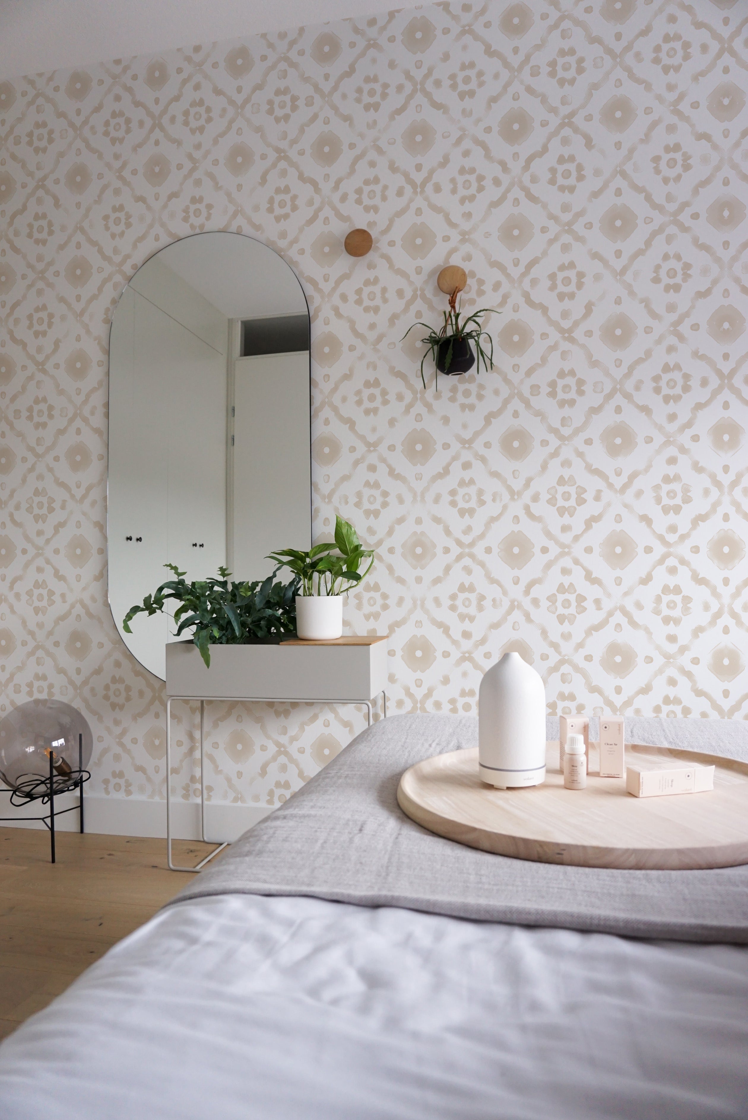 A cozy bedroom setting enhanced by Euclid Wallpaper, which covers the wall behind a bed and a small shelf with a decorative mirror and green houseplants. The wallpaper's soft beige geometric patterns on white provide a calm and inviting backdrop, complementing the room’s relaxed atmosphere.