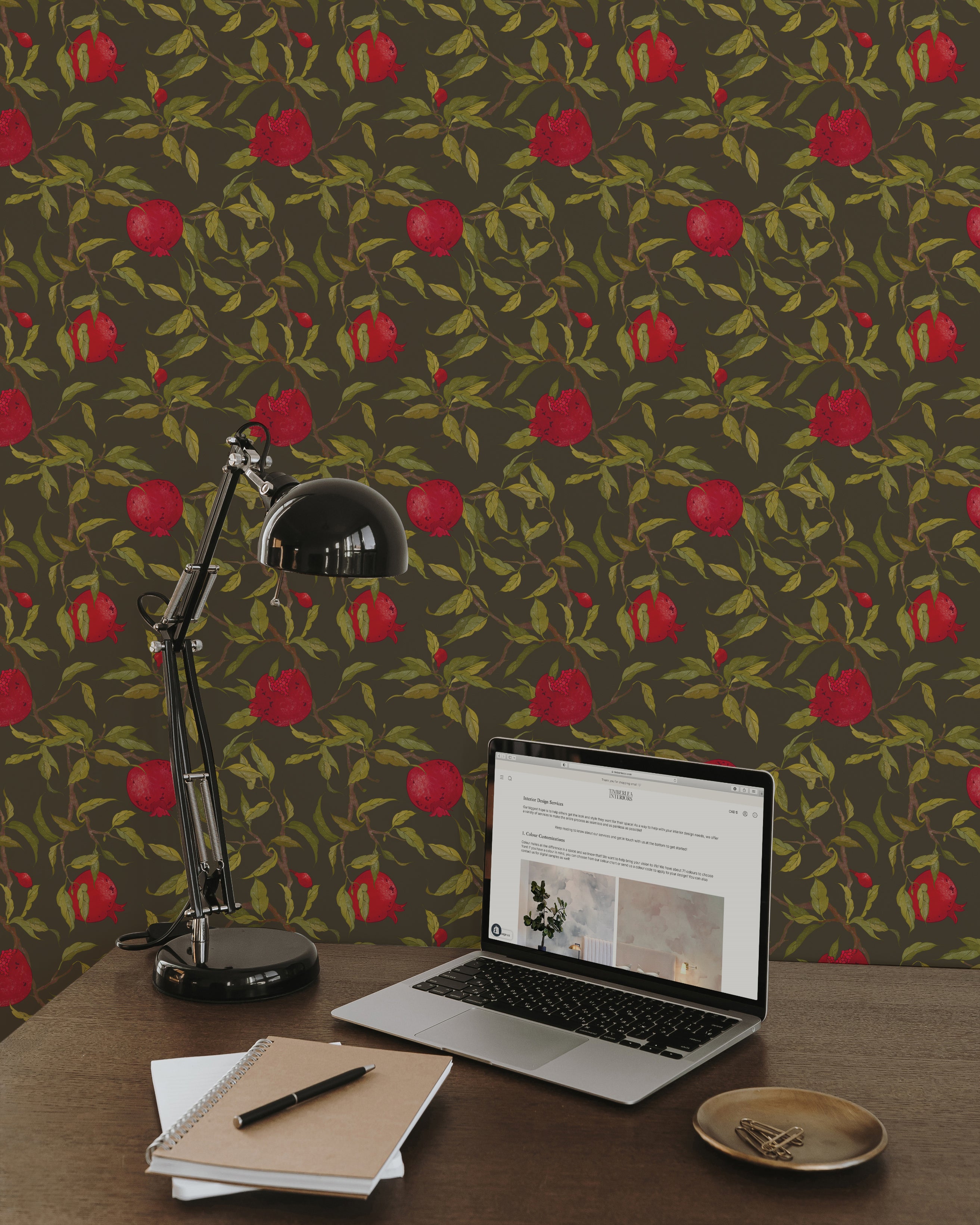An office setup with a sleek, black desk lamp illuminating a laptop on a wooden desk, set against a background of Vintage Pomegranate Wallpaper. The wallpaper’s vivid red pomegranates and green leaves on a dark backdrop add a touch of nature-inspired elegance to the workspace.