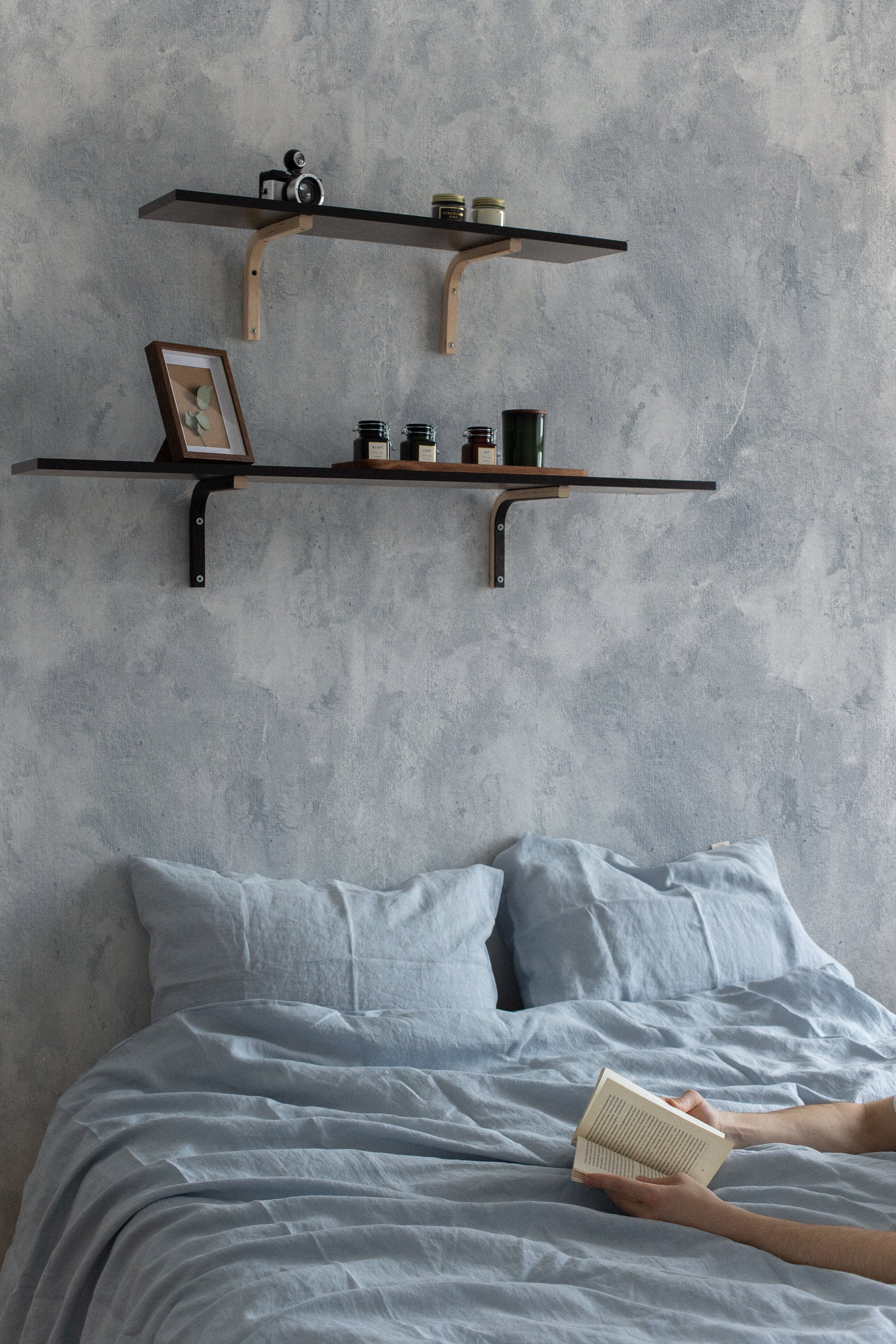 a bedroom scene where the Blue Limewash Wallpaper is applied on the wall behind a bed. The wallpaper's muted blue tones complement the linen bedding, enhancing the relaxed ambiance of the room. Two wooden shelves mounted on the wall display a simple arrangement of picture frames and jars, reinforcing the minimalist and serene aesthetic.