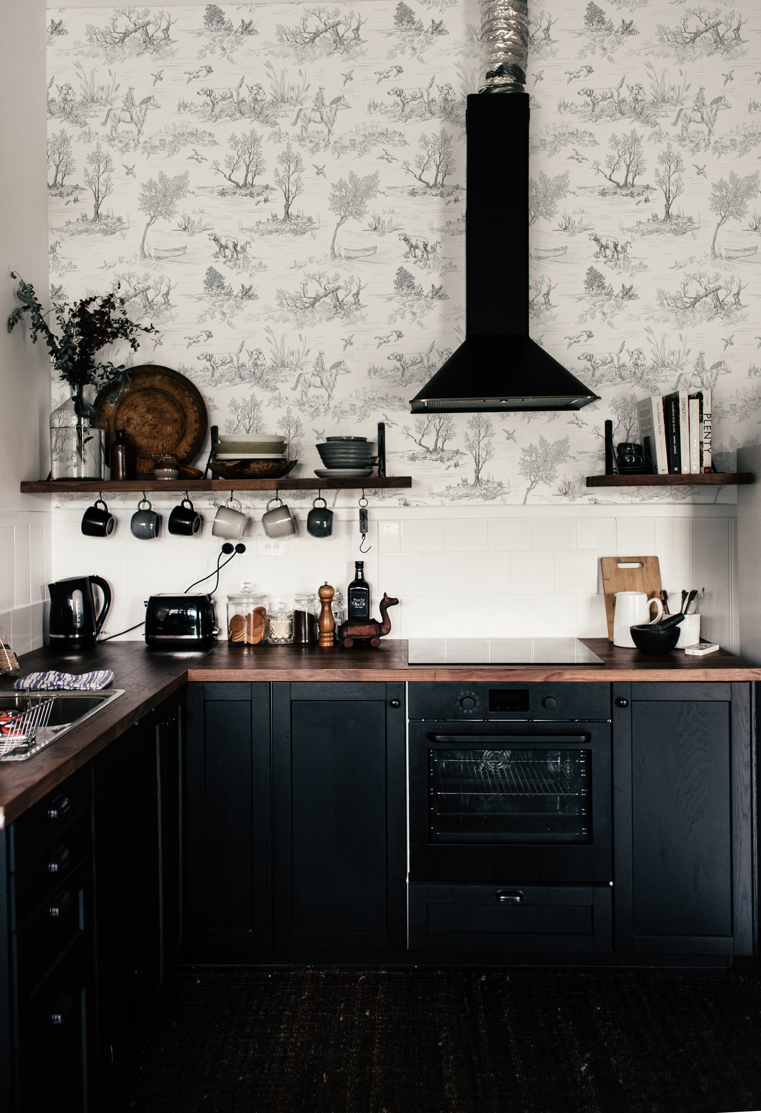 a kitchen scene with "Outdoor Friend Sketch Wallpaper." The wallpaper features a monochromatic sketch of a countryside scene with trees, dogs, and horses, creating a rustic and charming ambiance. The wall is complemented by dark cabinetry, a white tiled backsplash, wooden countertops, and a selection of kitchen utensils, contributing to the warm, traditional feel of the space.