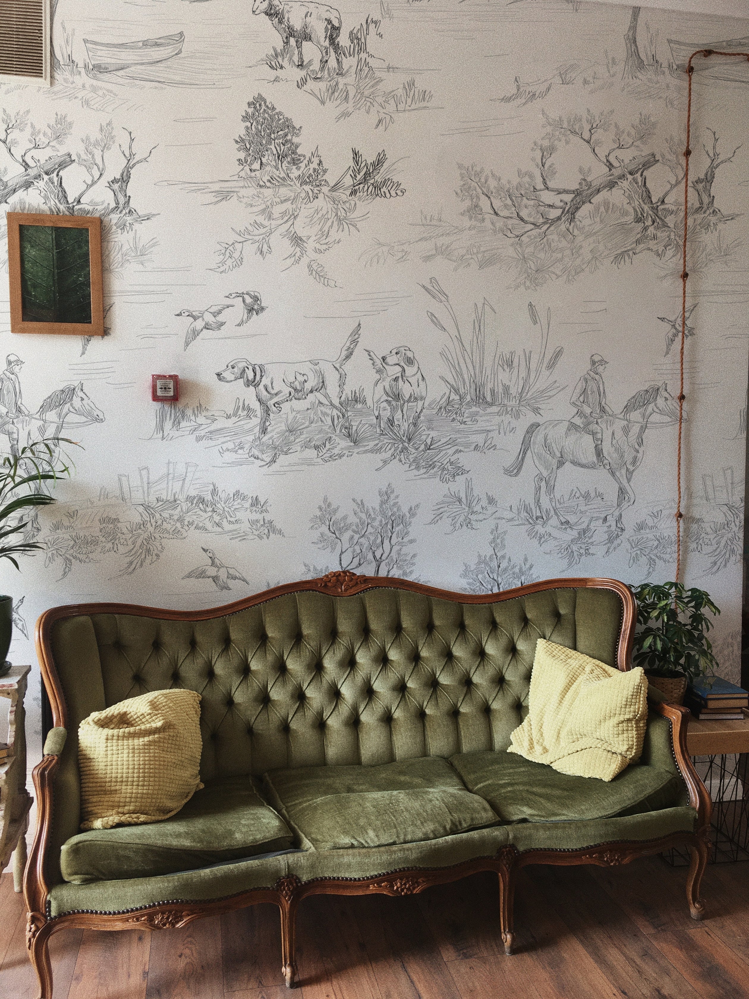 An elegant living room space highlighted by the Outdoor Friend Sketch Wallpaper. A vintage green velvet sofa with tufted backrest is set against the detailed sketch wallpaper, showing scenes of nature and outdoor activities, complemented by bright yellow pillows and indoor plants