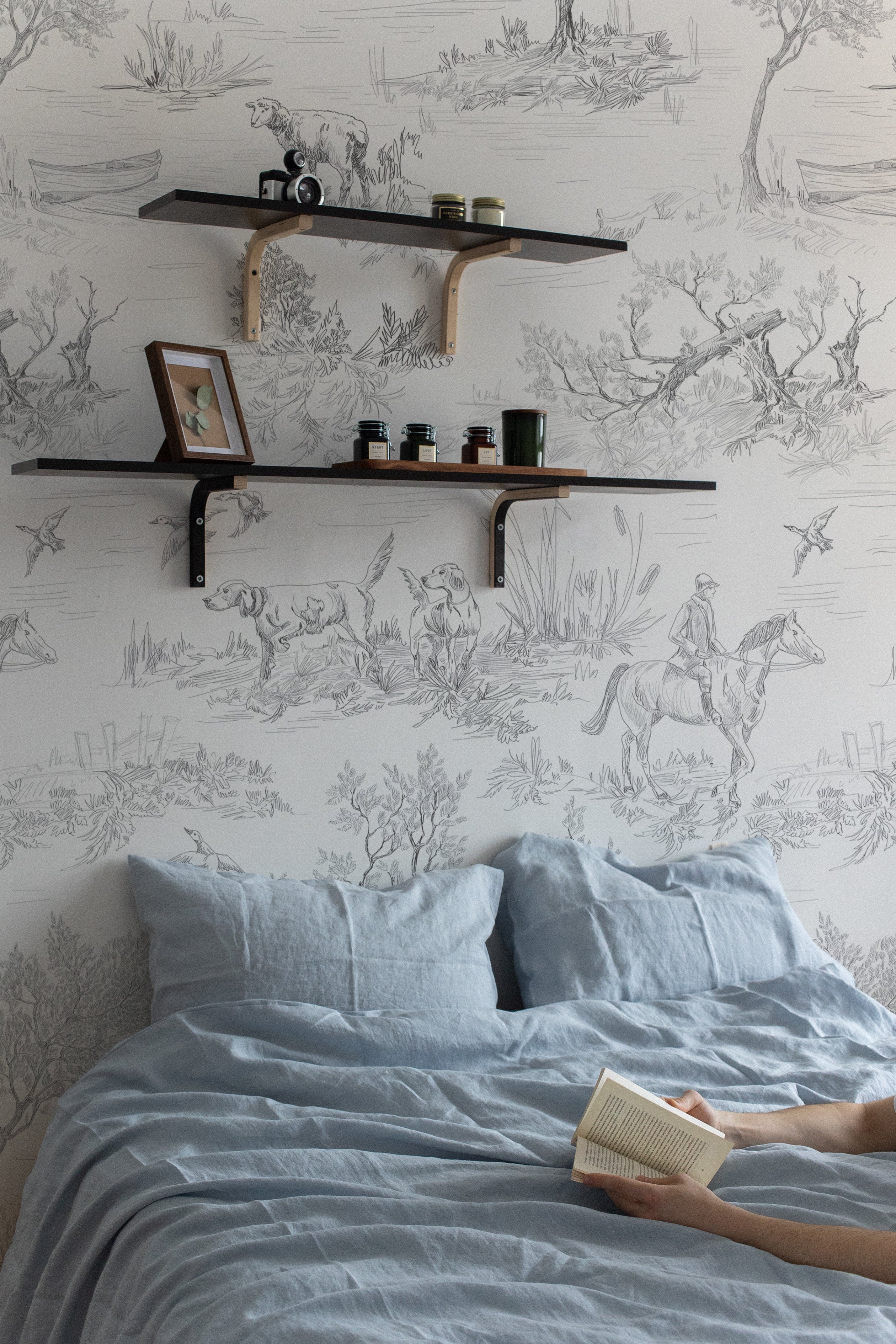 A cozy bedroom setting with Outdoor Friend Sketch Wallpaper. The wall behind the bed is adorned with intricate sketches depicting outdoor scenes with animals and hunters, providing a tranquil and artistic backdrop to the simple blue bedding and person reading a book.