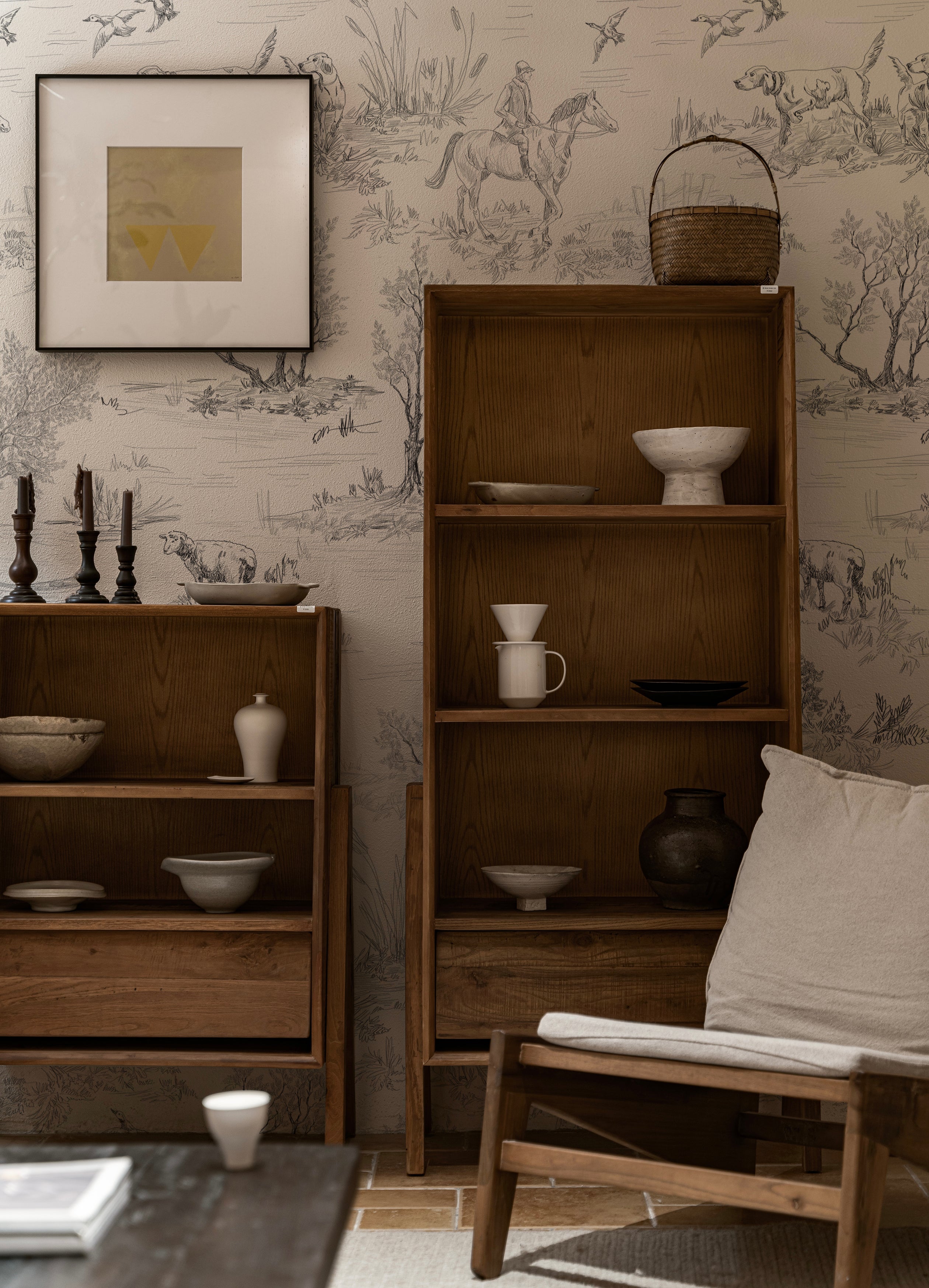 A rustic wooden bookcase against a backdrop of Outdoor Friend Sketch Wallpaper, featuring detailed sketches of outdoor scenes with hunters, dogs, and wildlife in a monochrome style. The decor includes various ceramic and wooden items, enhancing the naturalistic theme