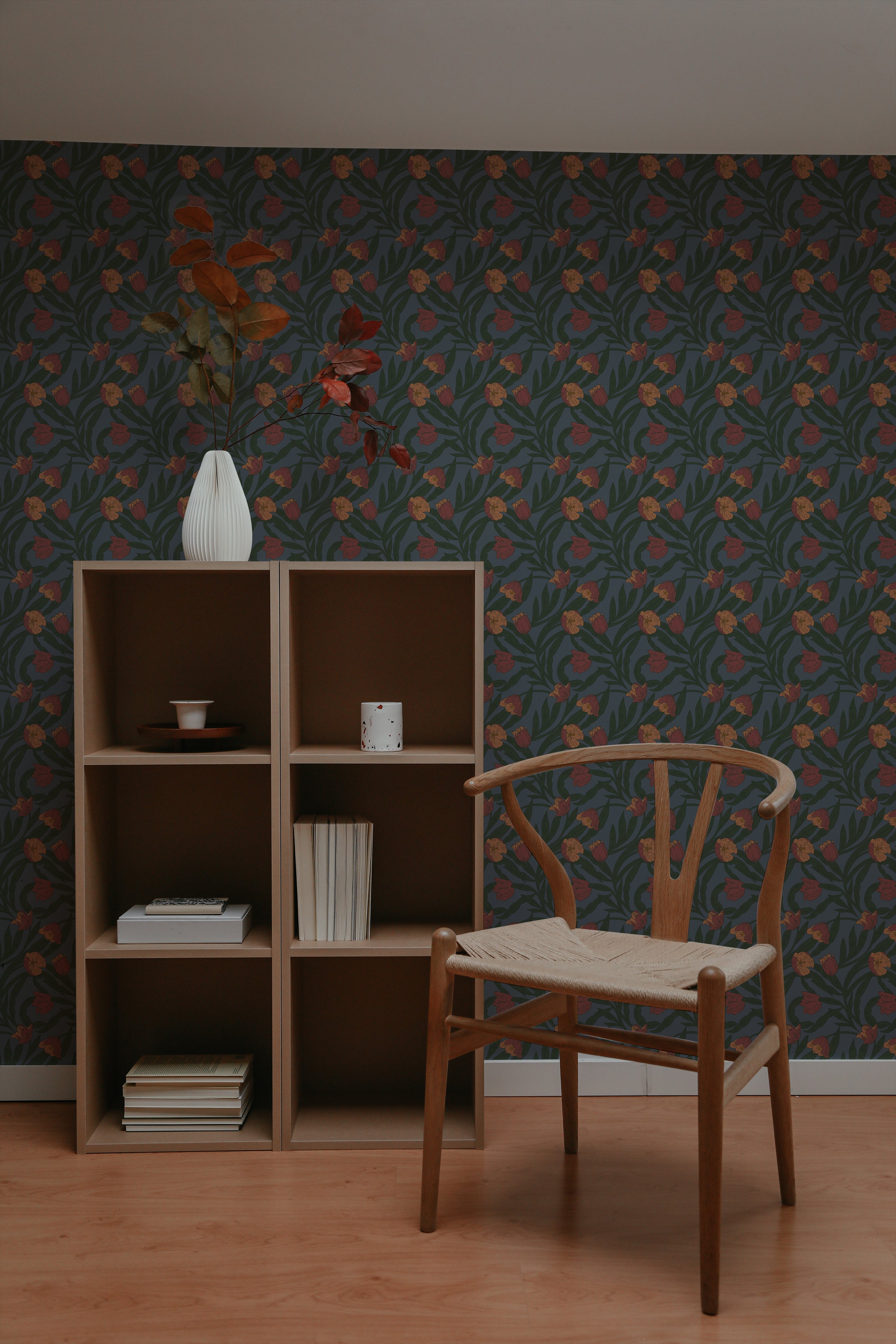 A serene reading nook with a classic wooden chair next to a matching bookshelf filled with books and decorative vases. The wall is covered in Morris Boho Wallpaper, showing a repetitive pattern of orange flowers and green leaves, creating a warm and inviting atmosphere.