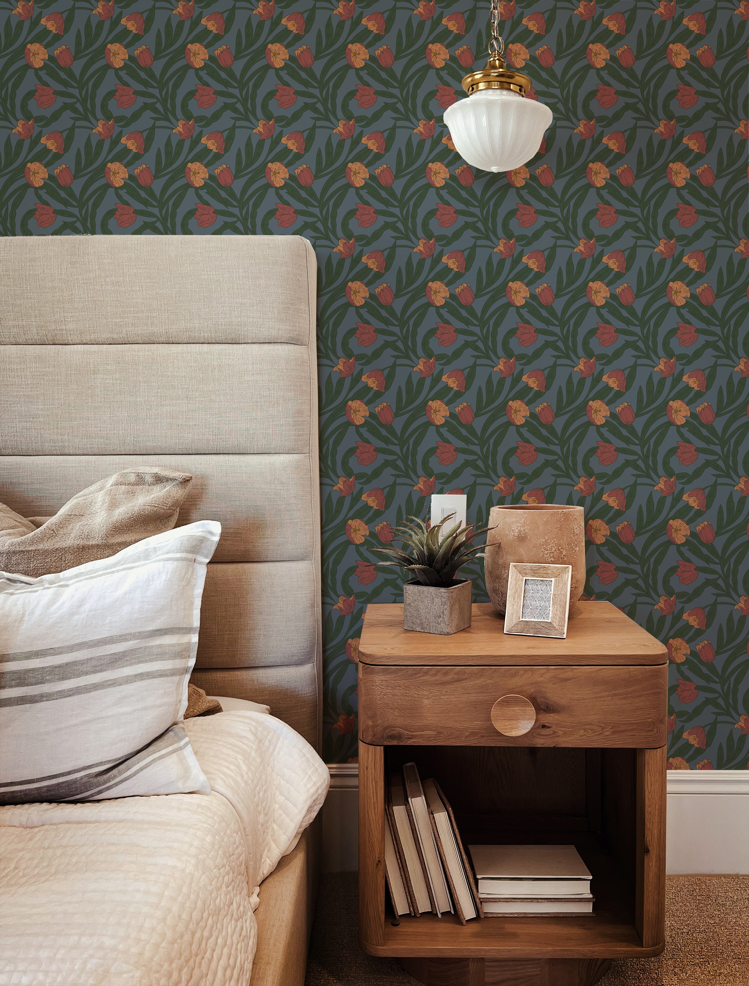 A stylish bedroom featuring a tall upholstered headboard and a modern bedside table with a small plant and decorative items. The room is highlighted by the Morris Boho Wallpaper, which has a pattern of bold orange flowers set against a dark green leafy background, adding a vibrant bohemian touch to the space.